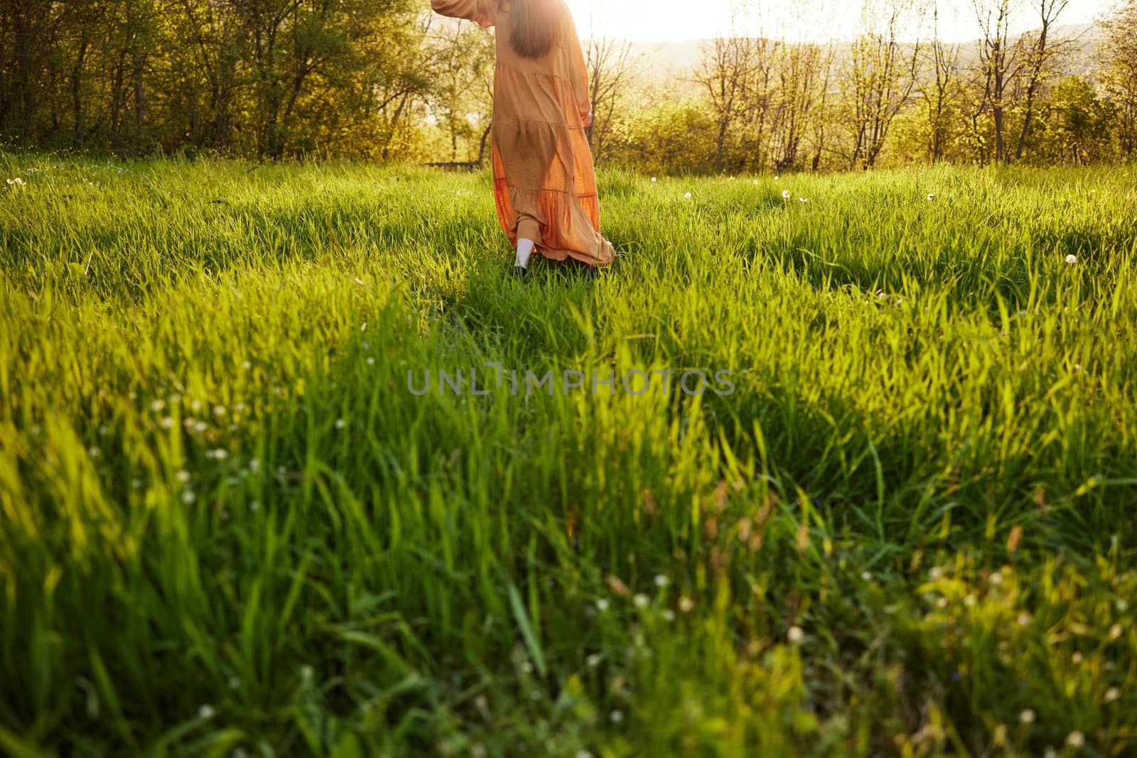 horizontal photo of a woman in an orange dress photographed without a face, standing in a green field during sunset, illuminated from the back. High quality photo