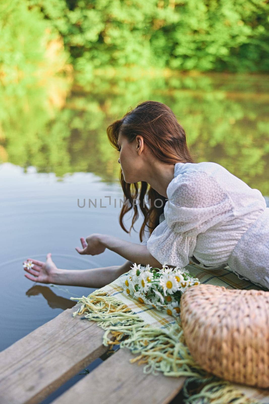 a woman in a light dress lying on a pier by the lake holds a chamomile flower in the water by Vichizh