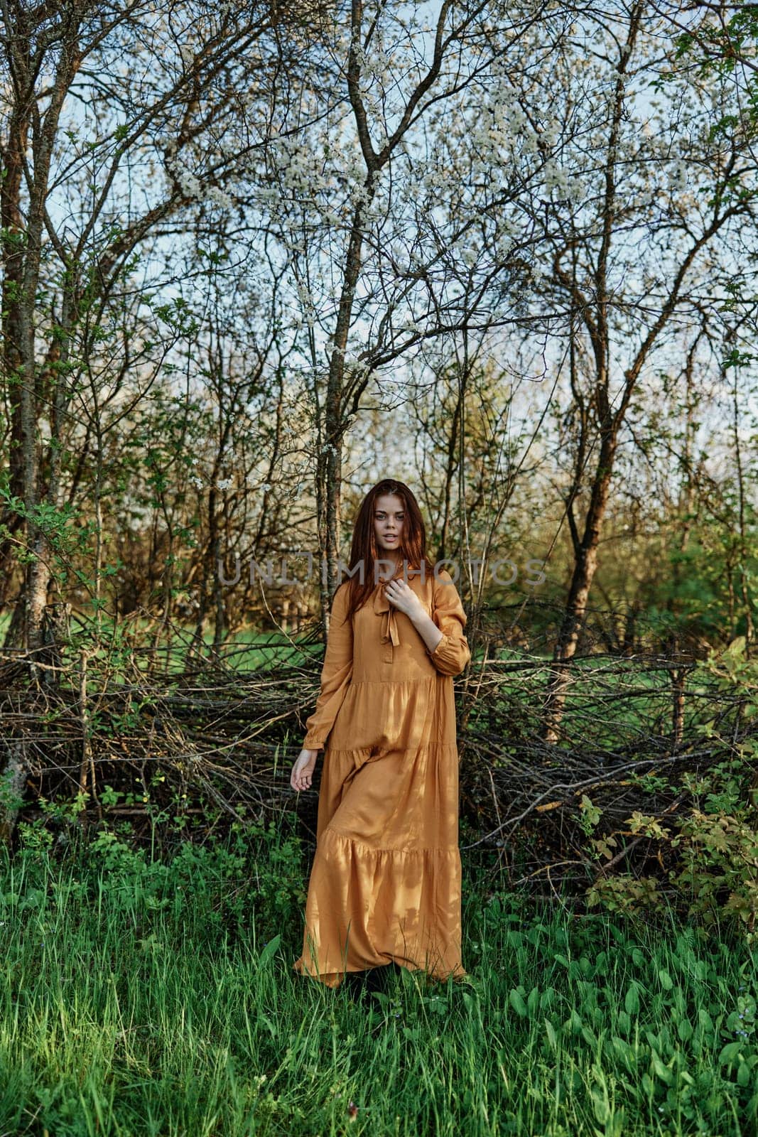 vertical photo of a beautiful, elegant woman in a stylish orange dress standing in the countryside in the shade of trees. High quality photo