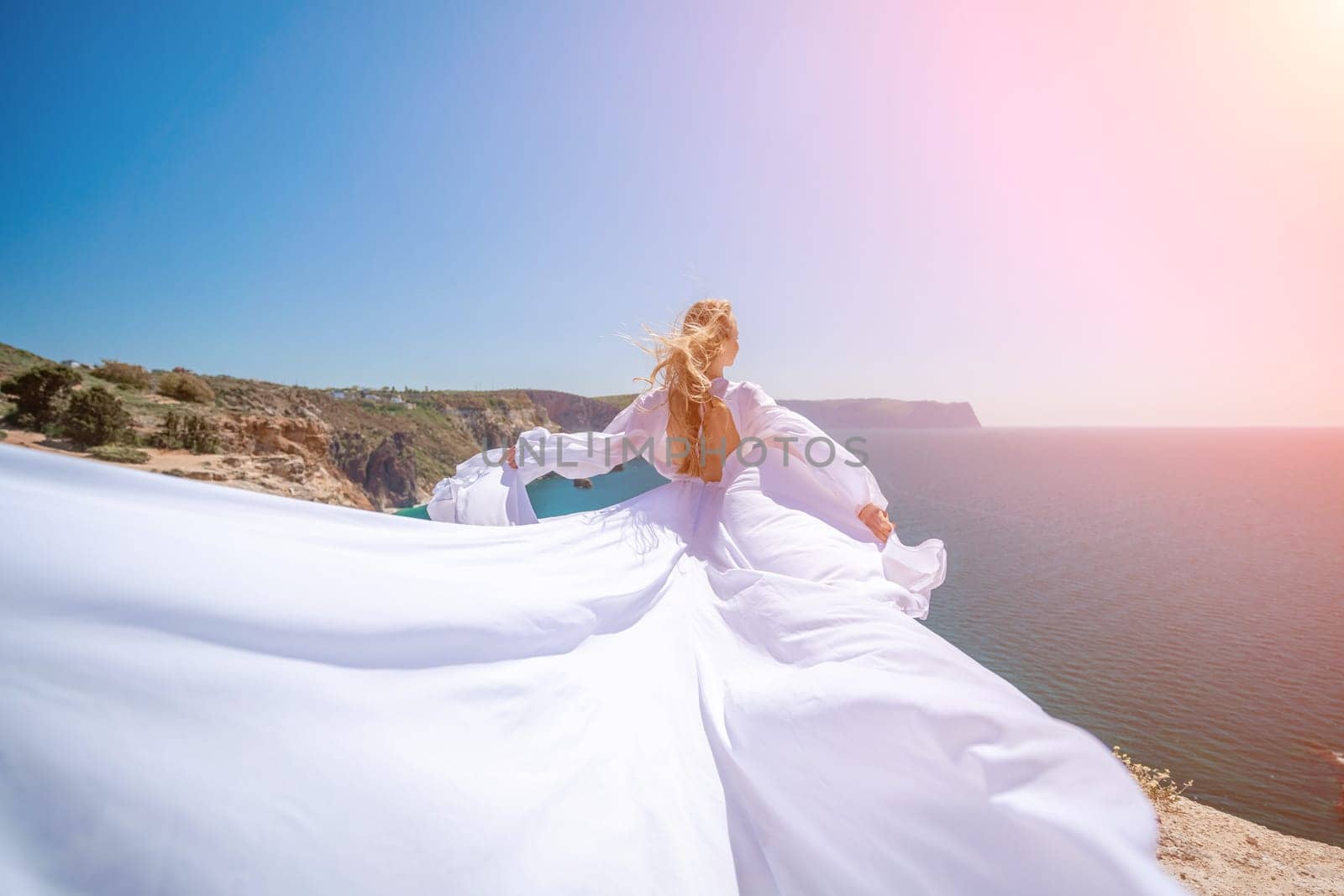 woman sea white dress. Blonde with long hair on a sunny seashore in a white flowing dress, rear view, silk fabric waving in the wind. Against the backdrop of the blue sky and mountains on the seashore