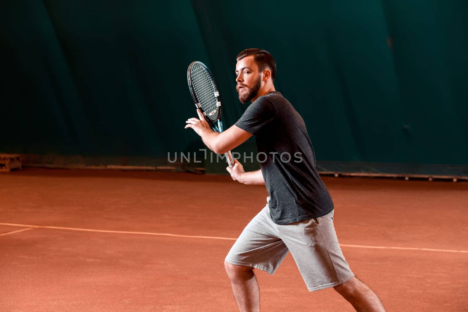 Handsome young man in t-shirt holding tennis racket and looking concentrated while standing on tennis court