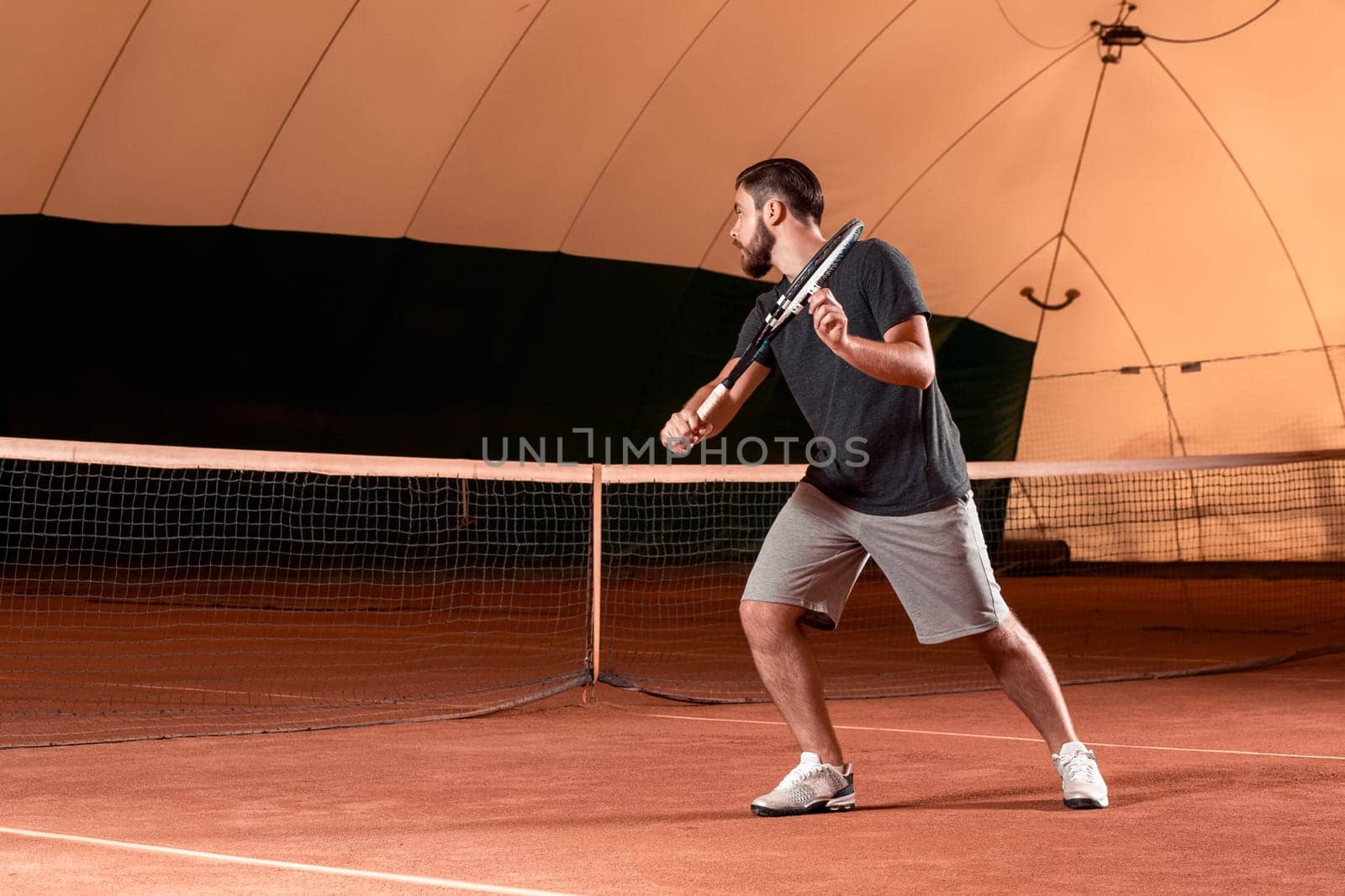 Handsome young man in t-shirt holding tennis racket and looking concentrated while standing on tennis court