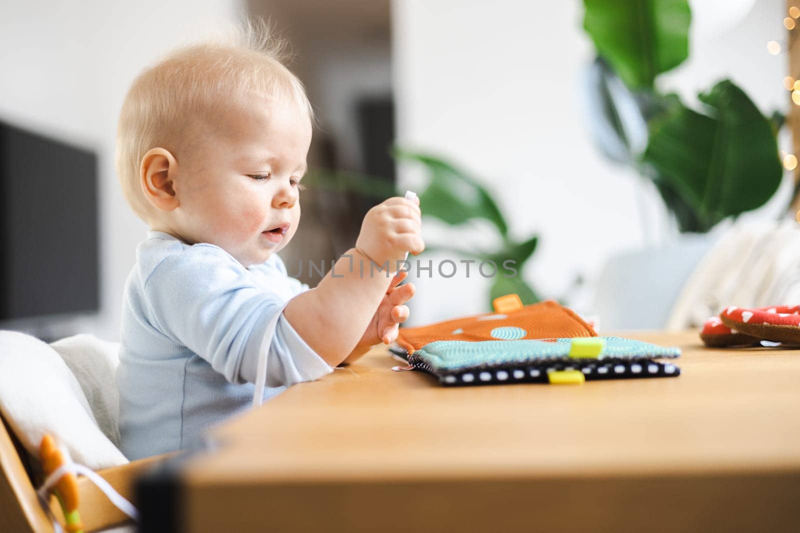 Happy infant sitting at dining table and playing with his toy in traditional scandinavian designer wooden high chair in modern bright atic home. Cute baby playing with toys by kasto
