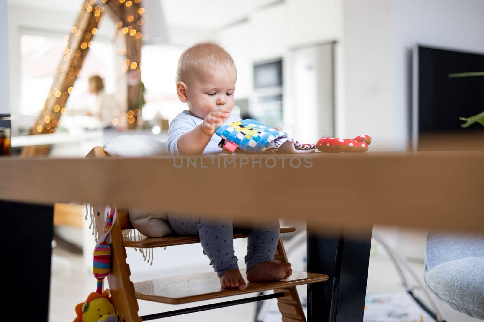 Happy infant sitting at dining table and playing with his toy in traditional scandinavian designer wooden high chair in modern bright atic home. Cute baby playing with toys.