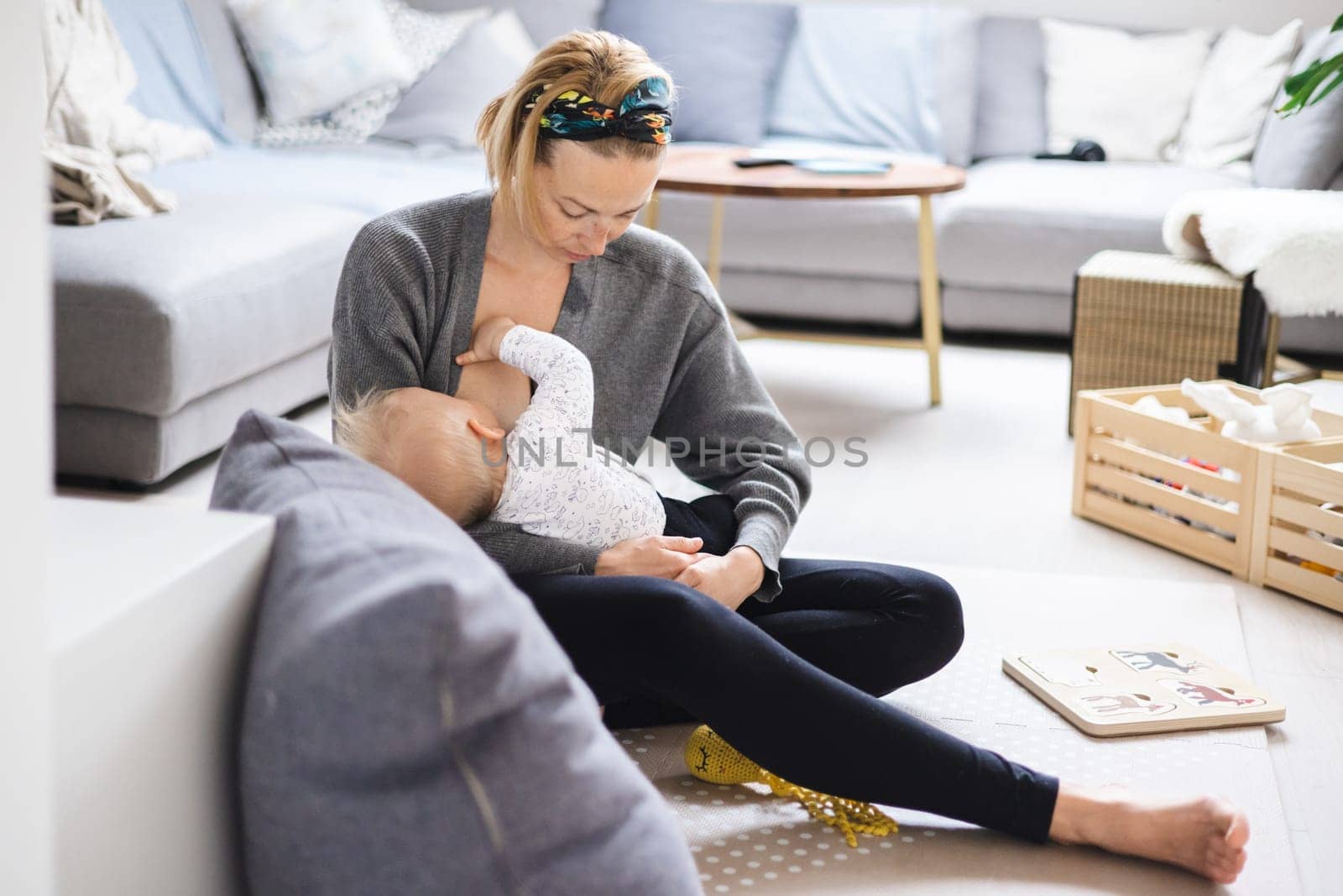 Young woman breastfeeding her infant baby boy casualy sitting on child's playing mat on living room floor at home