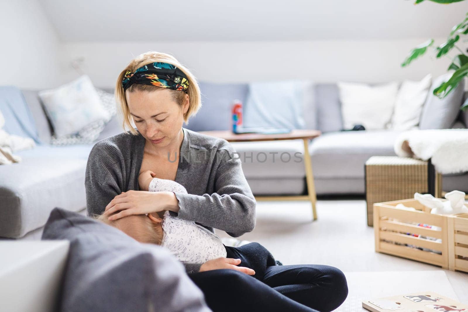 Young woman breastfeeding her infant baby boy casualy sitting on child's playing mat on living room floor at home