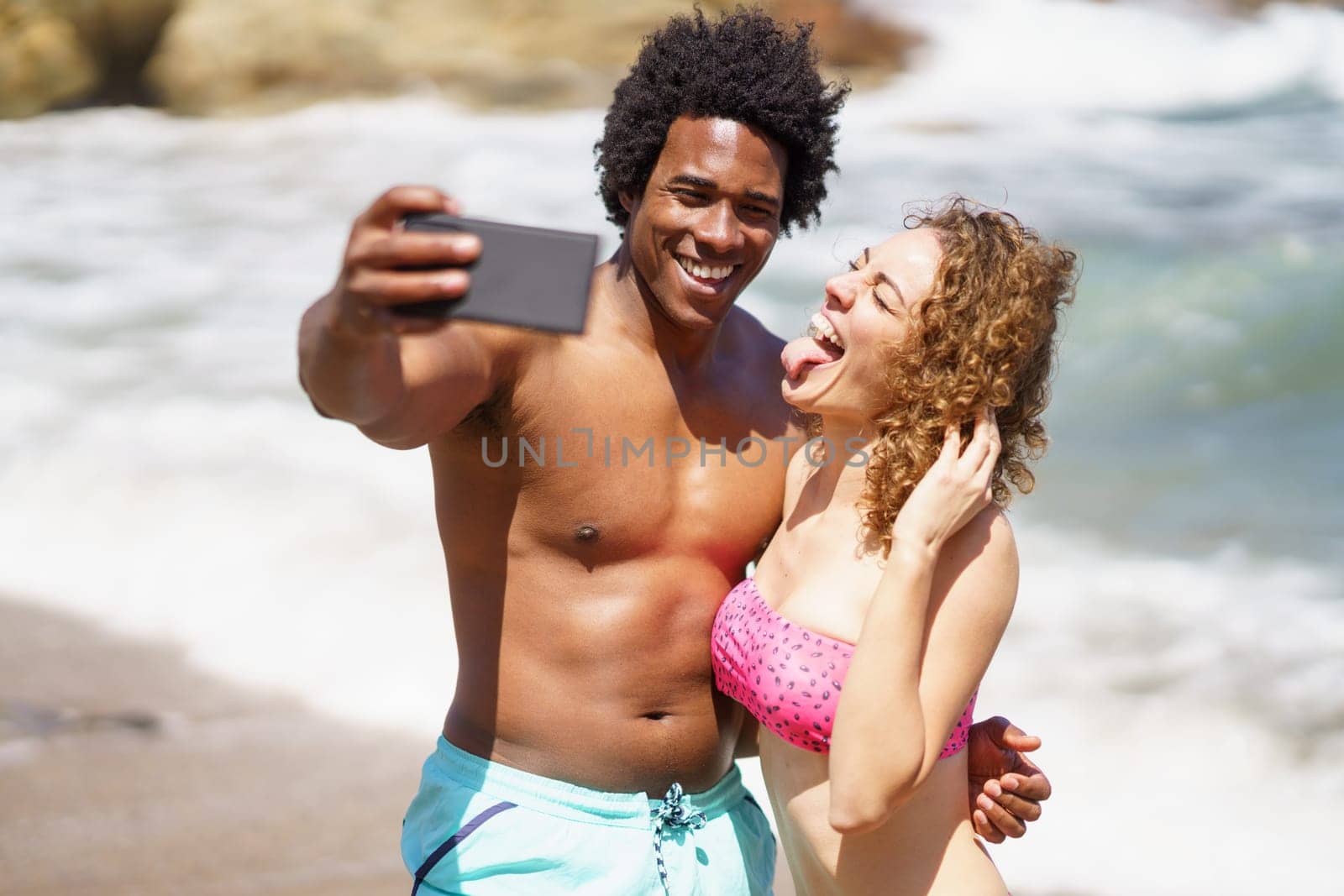 Happy African American man with naked torso and curly hair taking selfie on cellphone with girlfriend showing tongue against waving sea