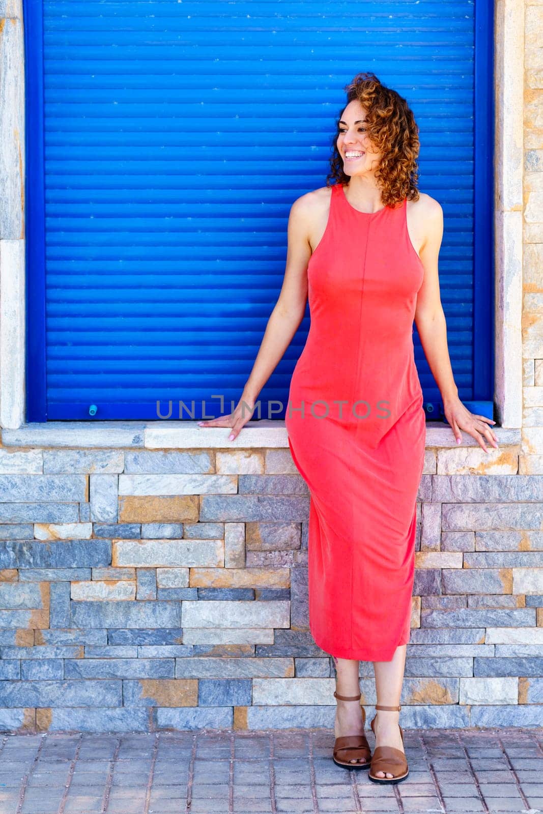 Full body of smiling female in red sundress and flat sandals looking away, while standing on paved street with hands on stone wall against closed blue shutters