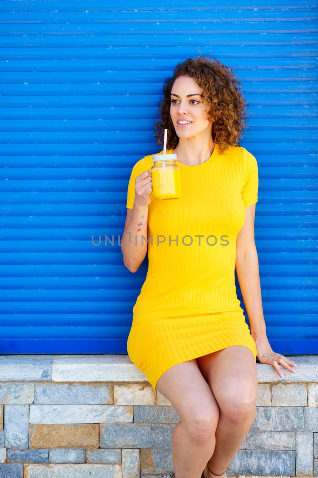 Cheerful young female in yellow dress with curly brown hair smiling and looking away while sitting on stone border with glass jar of fresh juice