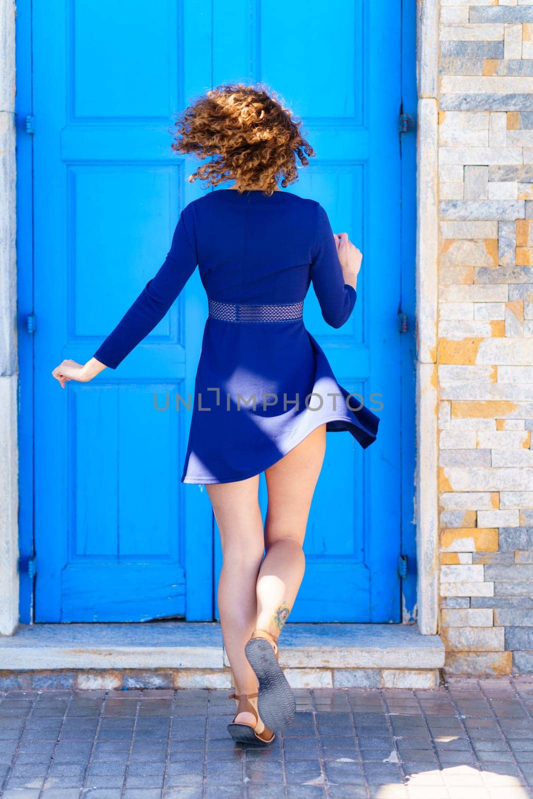 Back view full body of anonymous female in blue dress walking on paved street near bright door and brick building