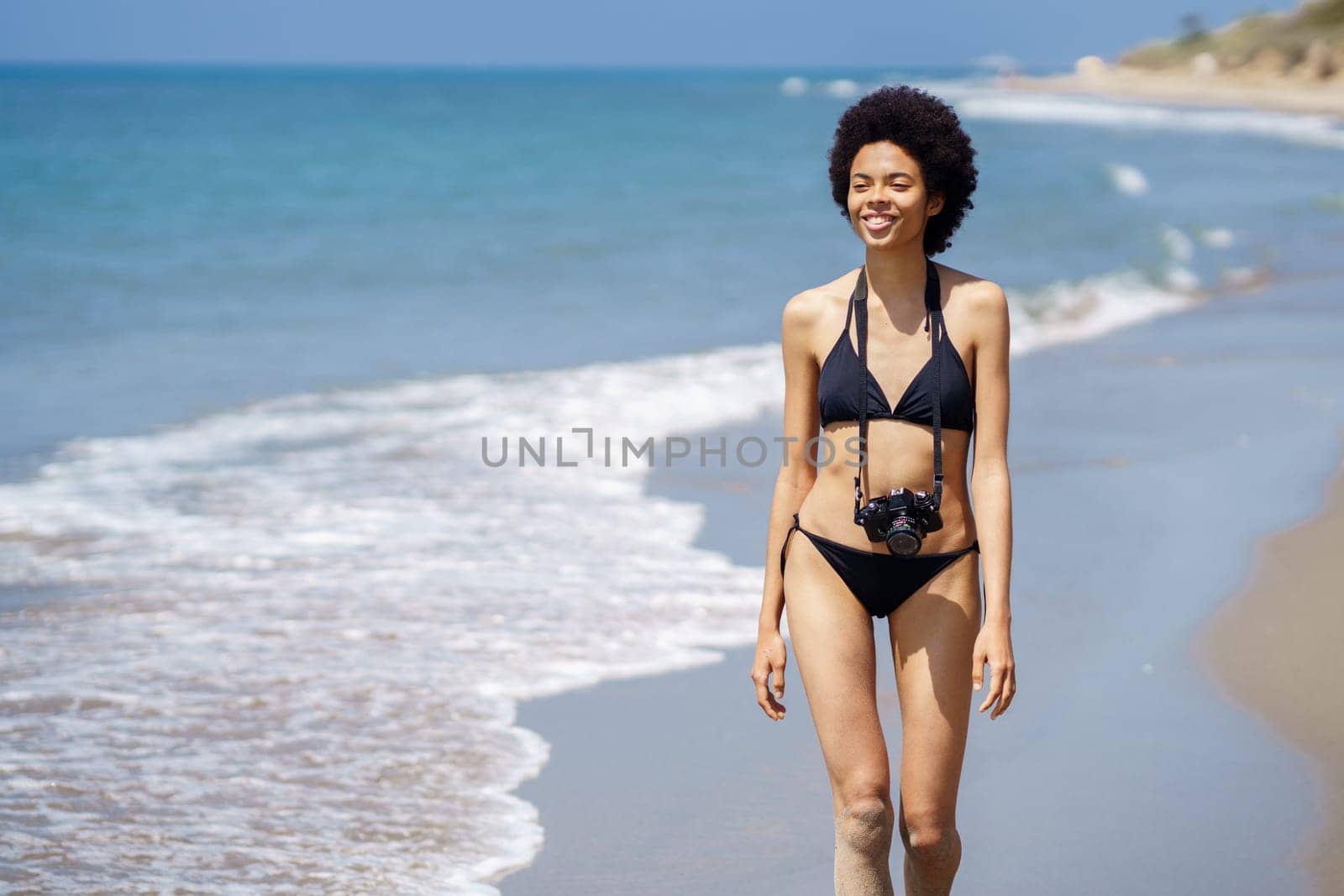 Cheerful African American female tourist in bikini with photo camera walking on wet shore near sea during trip in tropical resort on summer day