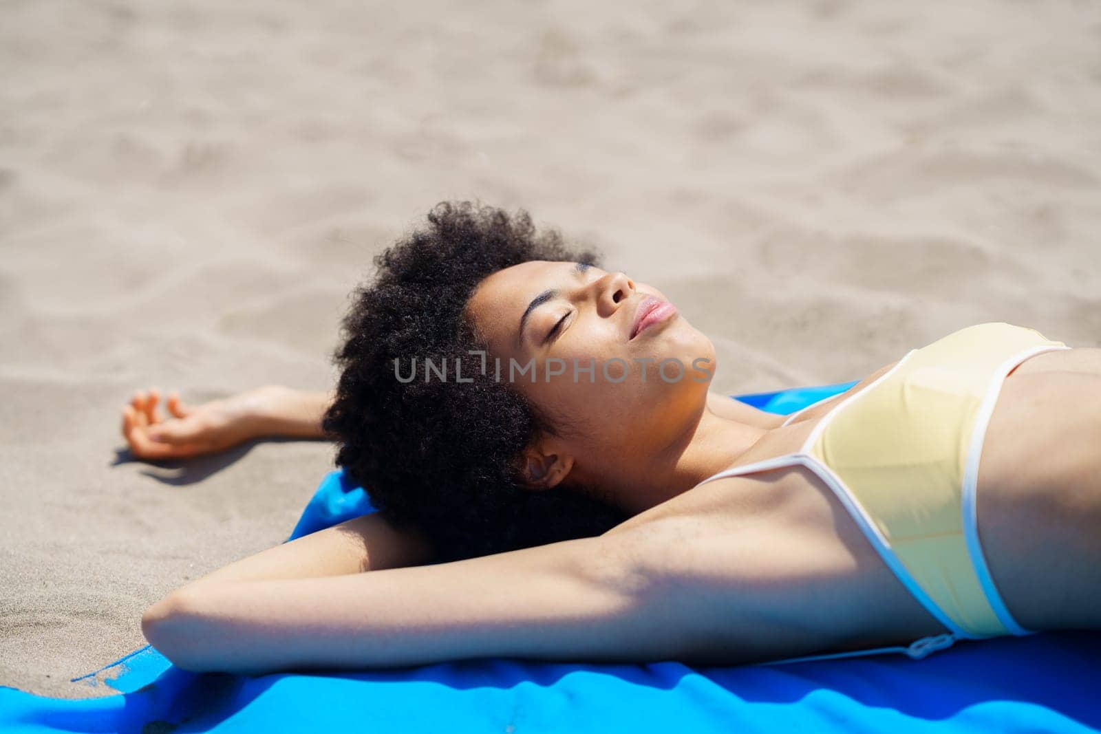 Happy young African American female tourist with curly hair in trendy bikini lying on sandy beach with closed eyes while sunbathing during summer holidays