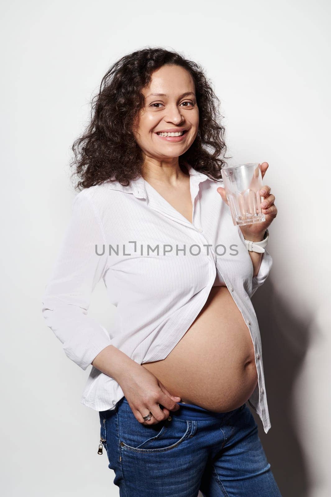 Vertical studio portrait of curly haired pretty brunette, happy smiling pregnant woman holding a glass of water, enjoying her carefree pregnancy, posing with her naked belly, white isolated background