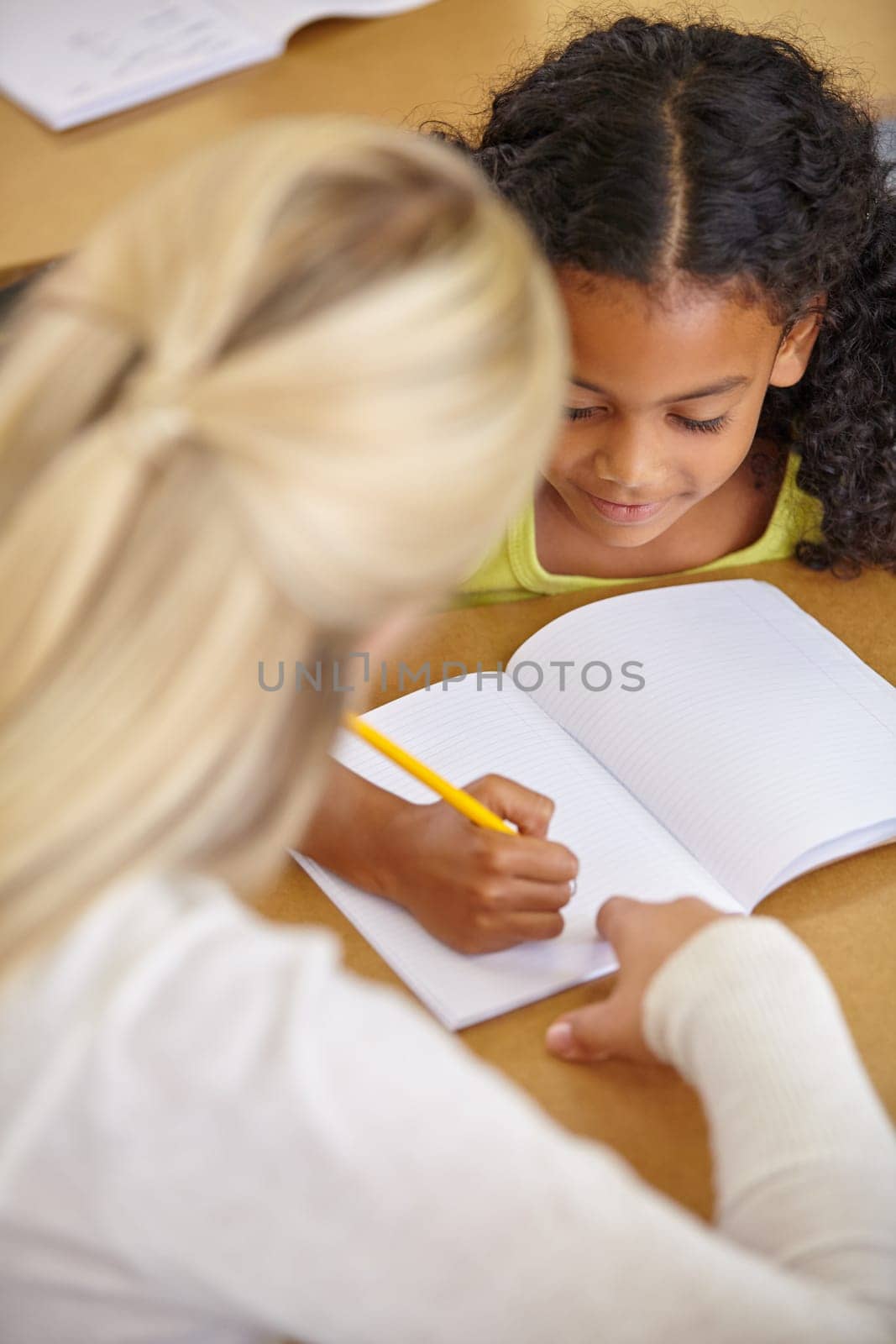 Teacher helping student in classroom, education and writing in notebook with pencil, teaching and learning for development. Scholarship, academic study and young girl and woman in lesson at school by YuriArcurs