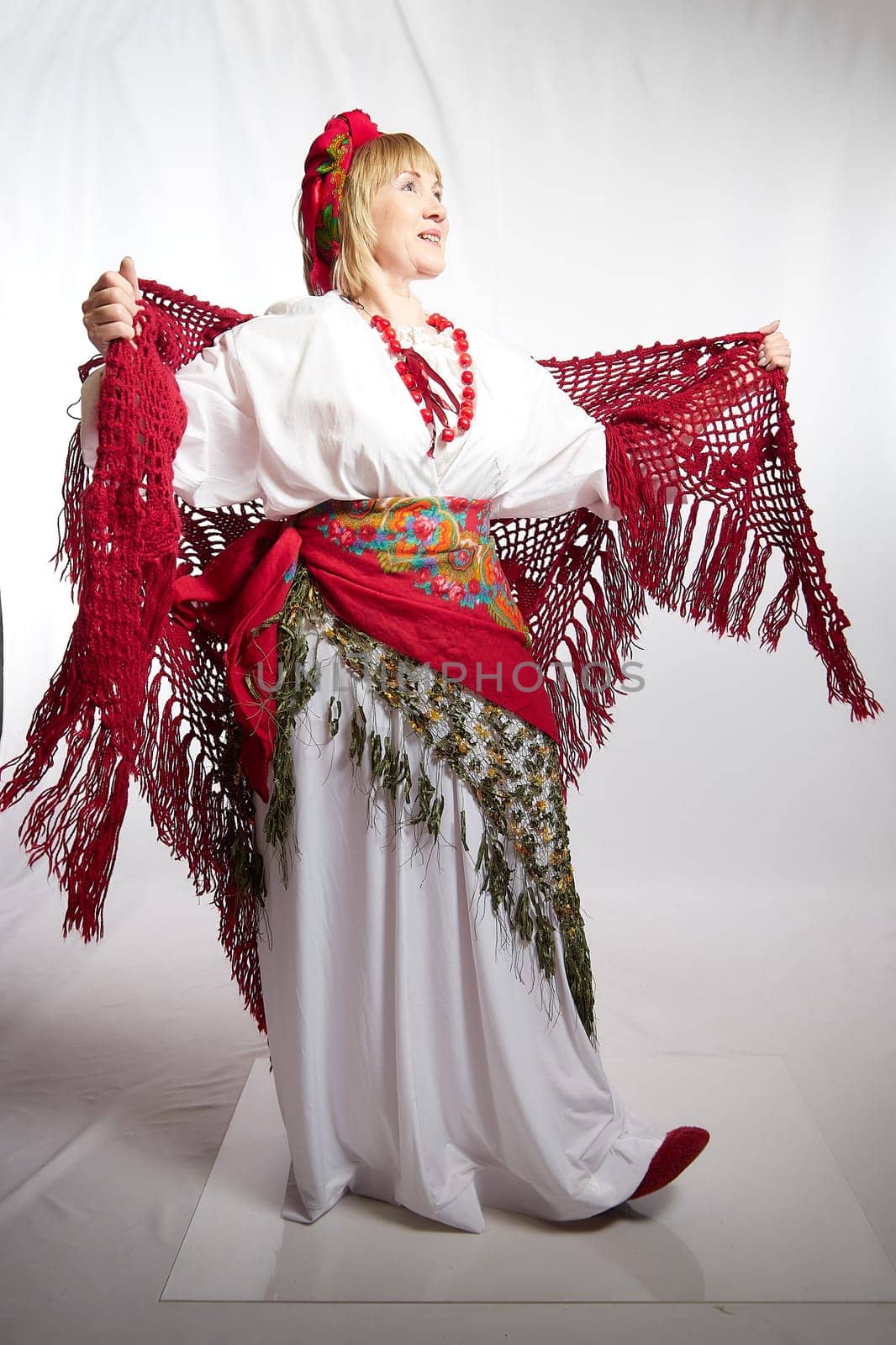 Cheerful funny adult mature woman solokha in a red knitted shawl. Female model in clothes of national ethnic Slavic style. Stylized Ukrainian, Belarusian or Russian woman poses in comic photo shoot