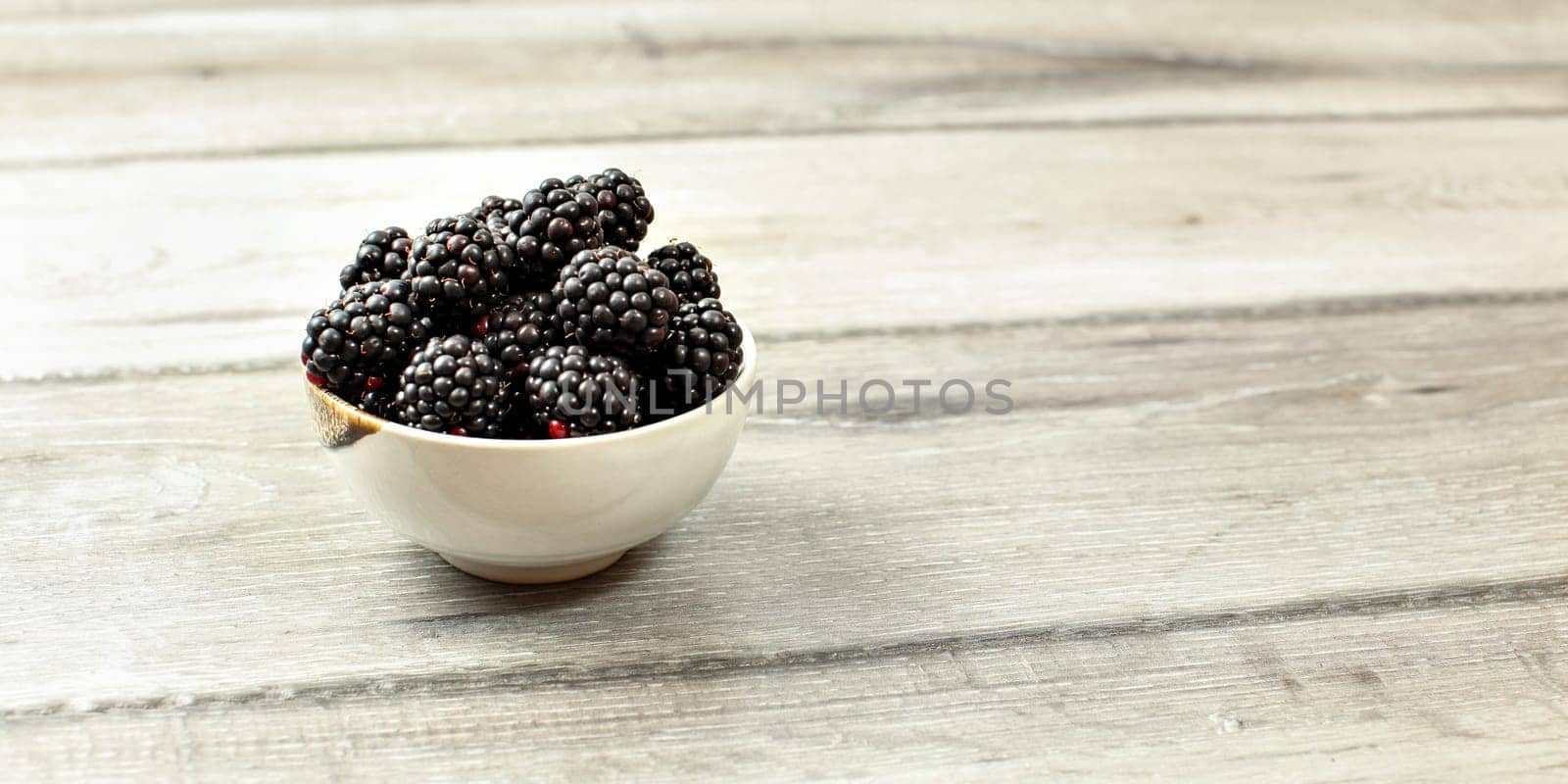 Small bowl with blackberries on wooden desk, space for text on right side.