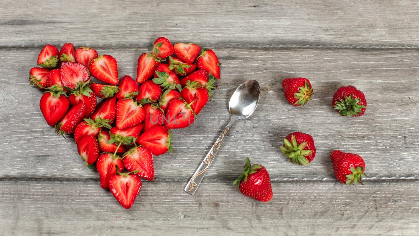 Top down view, strawberry pieces arranged in shape of heart, spoon and some spilled strawberries near, on gray wood desk.