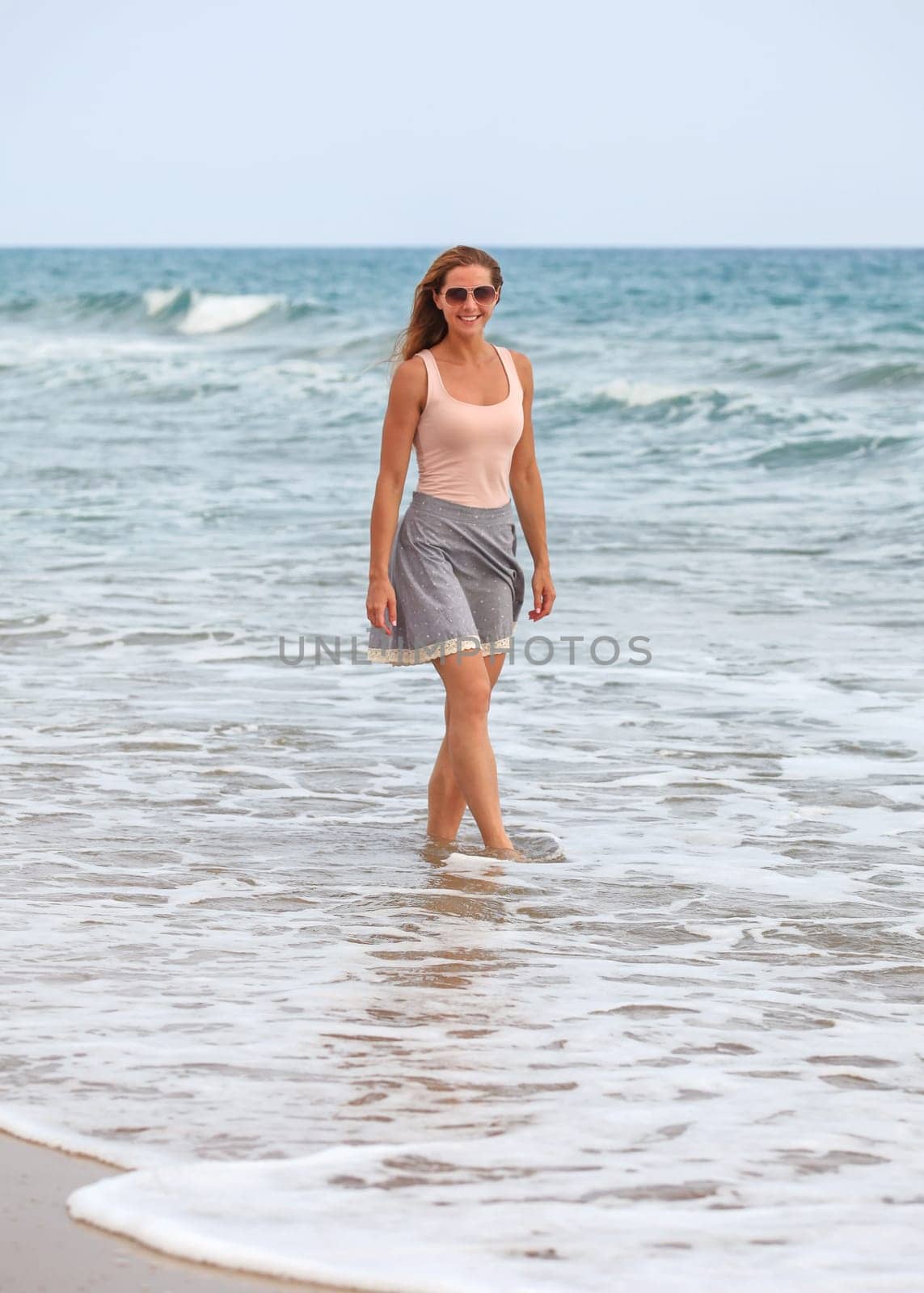 Young woman in skirt, t shirt and sunglasses standing in shallow water on the beach during overcast day, sea behind her.