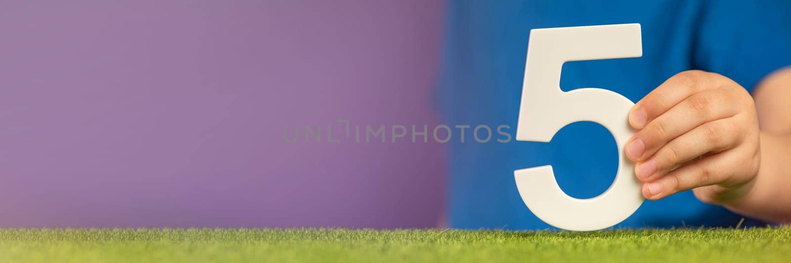Number 5 in hand. A hand holds a white number five on green grass and a purple background, five percent per annum. Concept of victory day in the Netherlands and Mexico, 5 May. by SERSOL