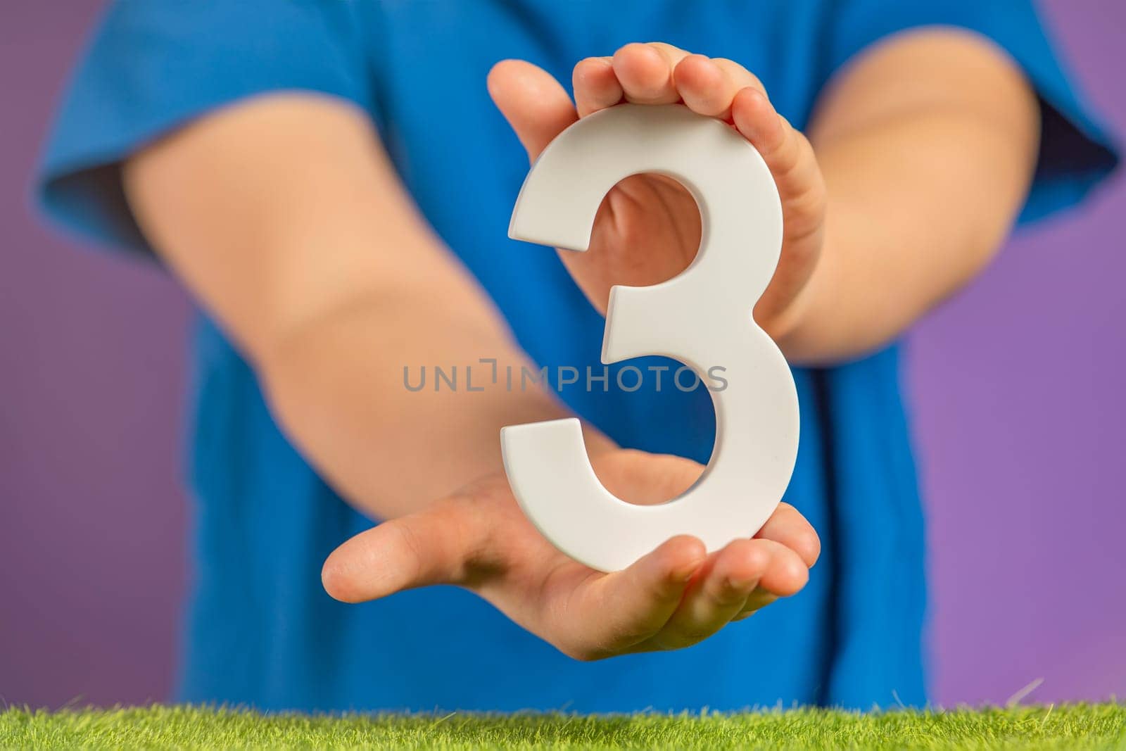 Number 3 in hand. Hand holding white number three over green grass and purple background, birthday concept, three steps to success, triple win. Banner with copy space. by SERSOL