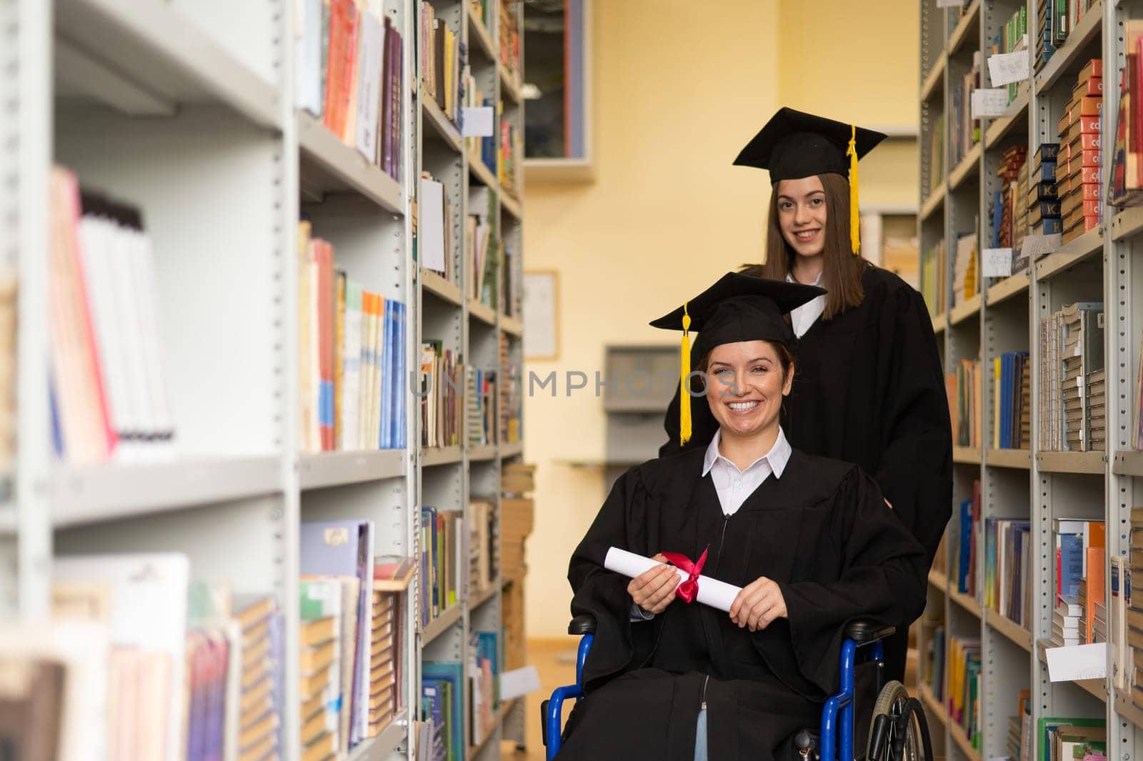Happy young woman and woman in wheelchair in graduate gown with diploma in hands in library. Inclusive education