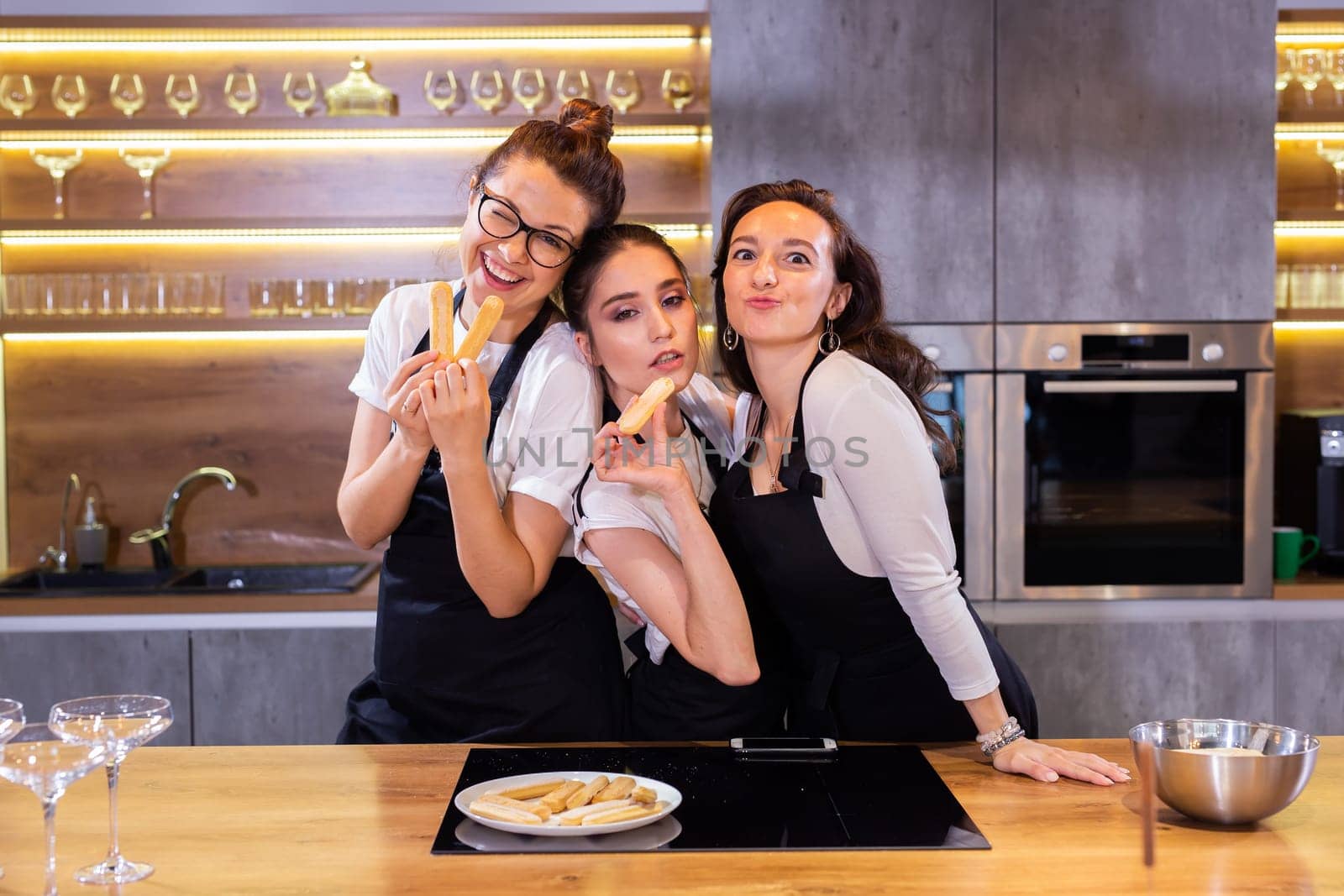 Three funny female chef in uniform holding cookies while smiling and having fun at camera in kitchen