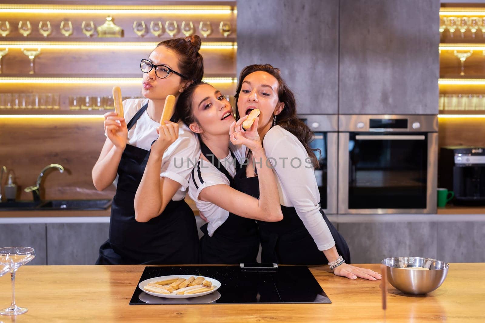 Three funny female chef in uniform holding cookies while smiling and having fun at camera in the kitchen by Satura86