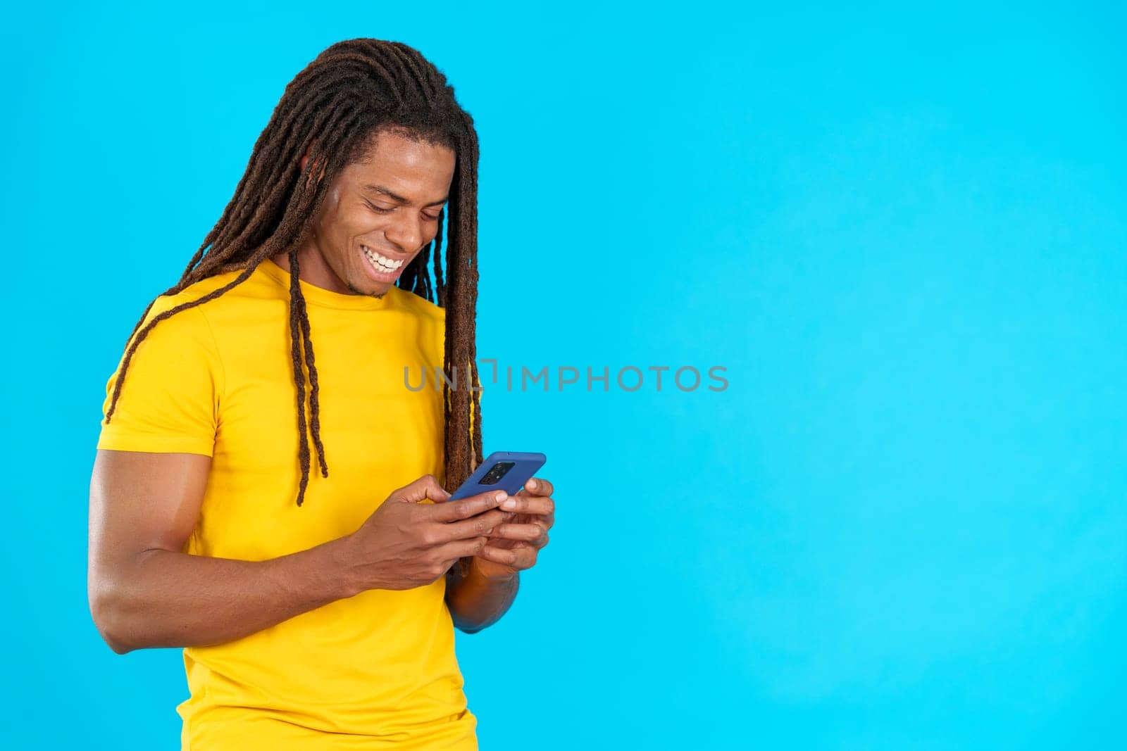 Happy latin man with dreadlocks smiling while using the mobile in studio with blue background