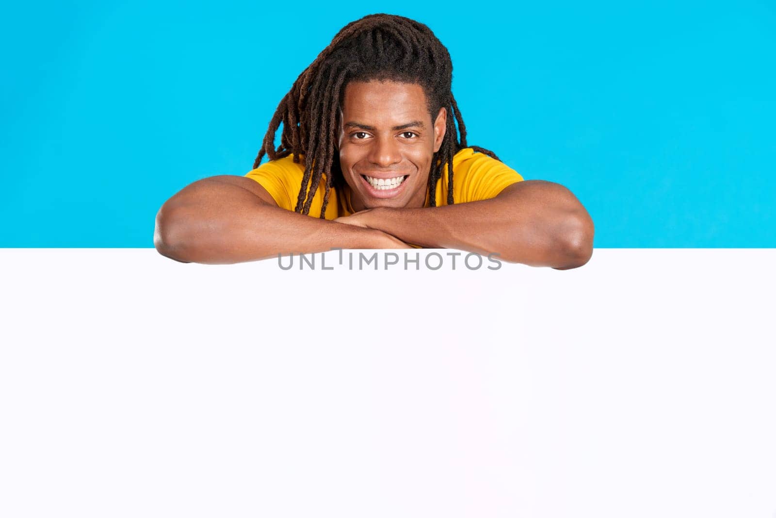 Latin man with dreadlocks leaning on a blank board in studio with blue background