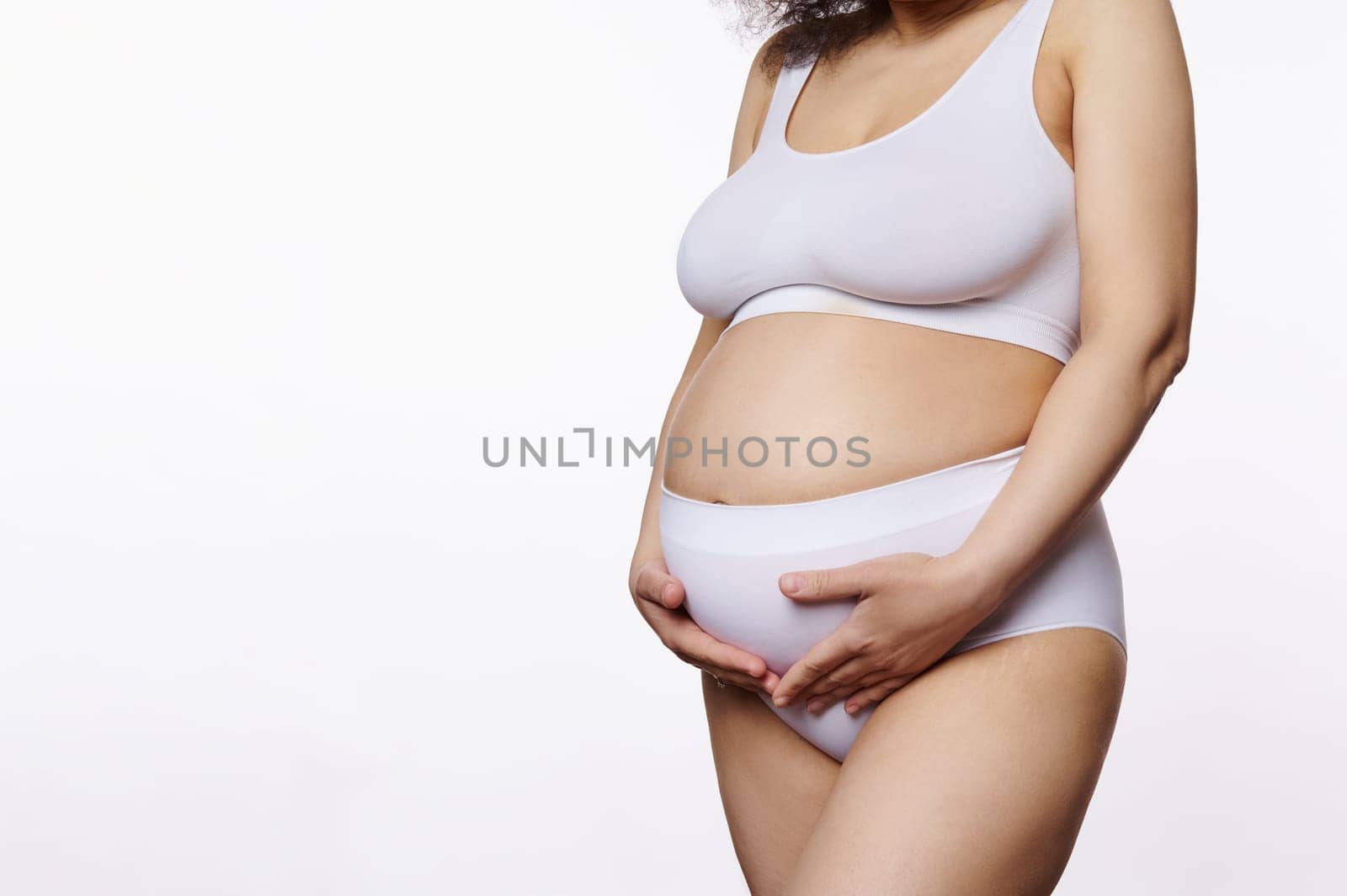 Close-up studio shot of a pregnant woman, holding her belly, posing in white lingerie over isolated background with copy ad space. Nudity. Body positivity. Natural beauty people. Pregnancy. Maternity