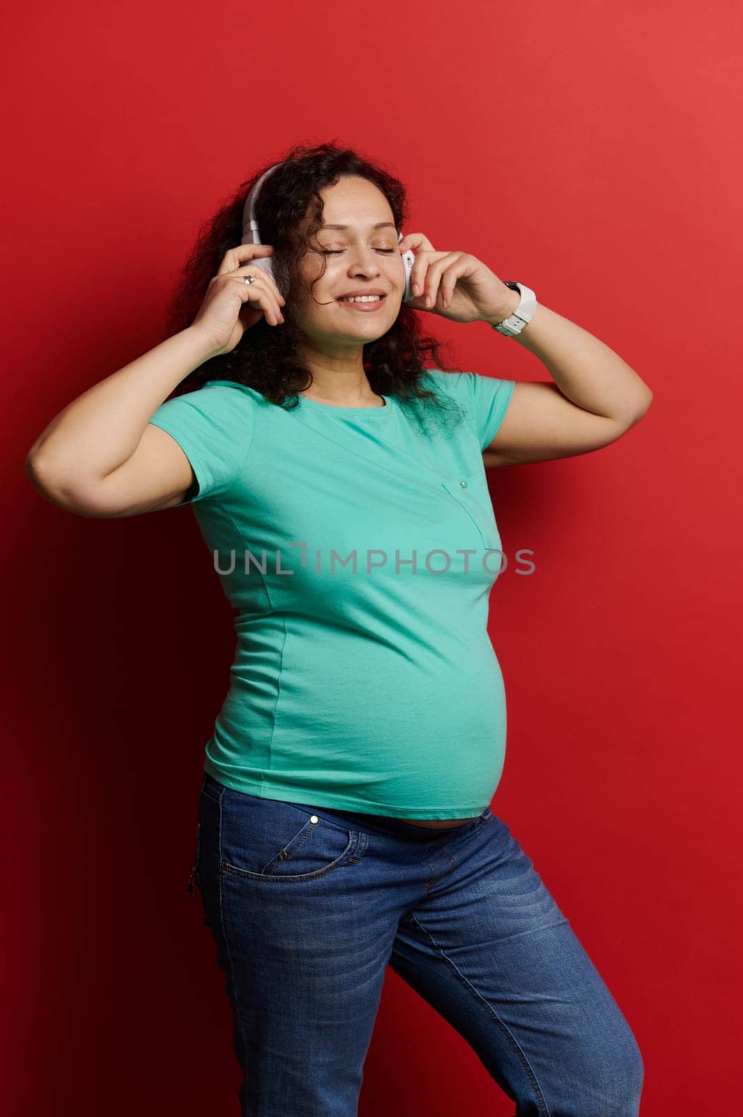 Portrait of a middle aged curly haired multi ethnic beautiful pregnant woman in turquoise t-shirt and blue jeans, enjoying soothing music, wearing wireless headphones, isolated on red background