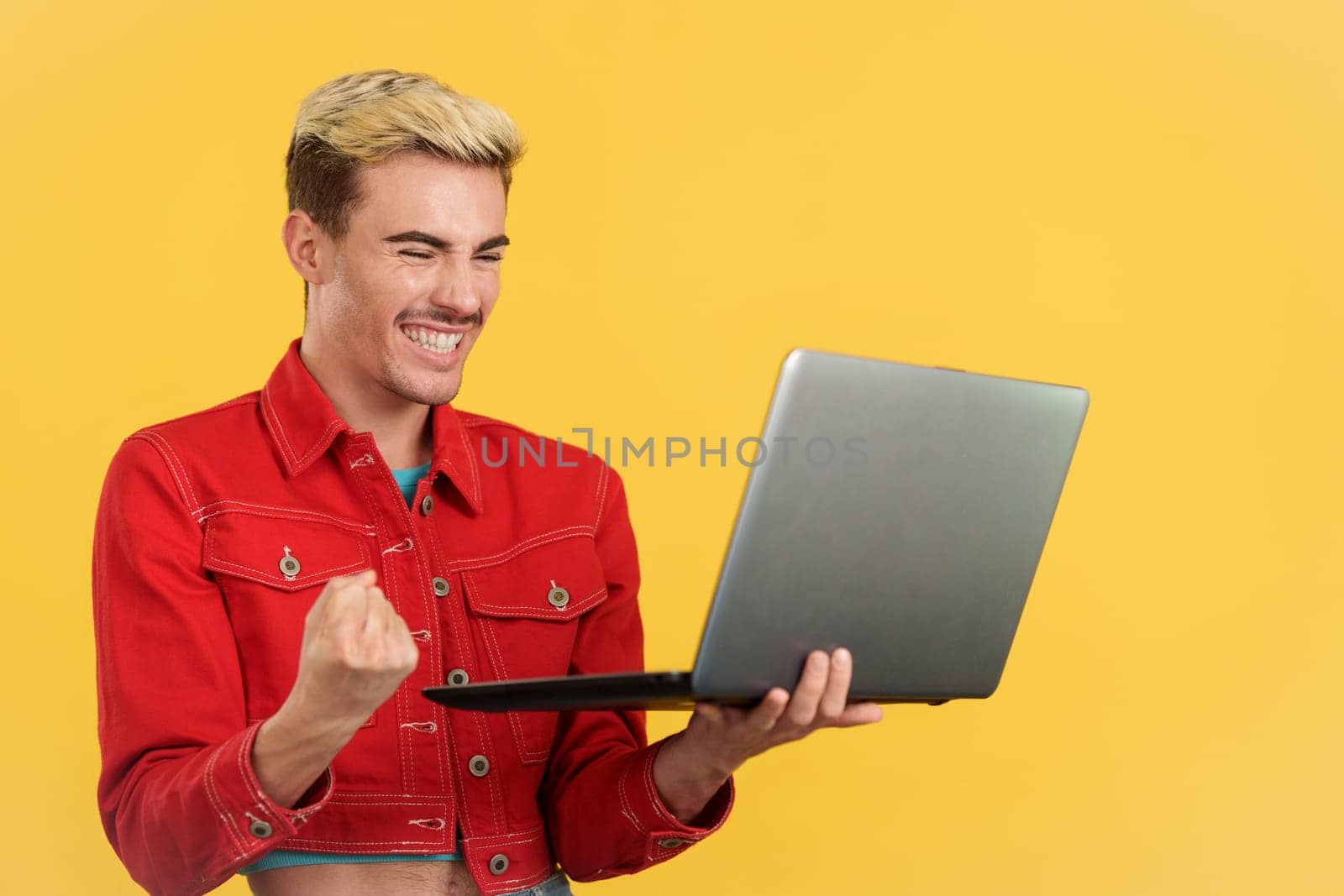 Gay man celebrating while looking at the laptop in studio with yellow background