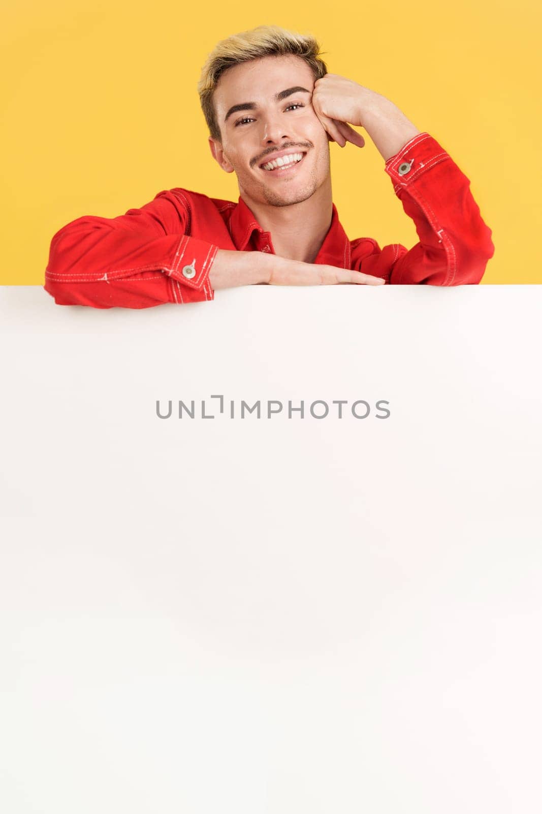 Cheerful gay man leaning on a blank board in studio with yellow background