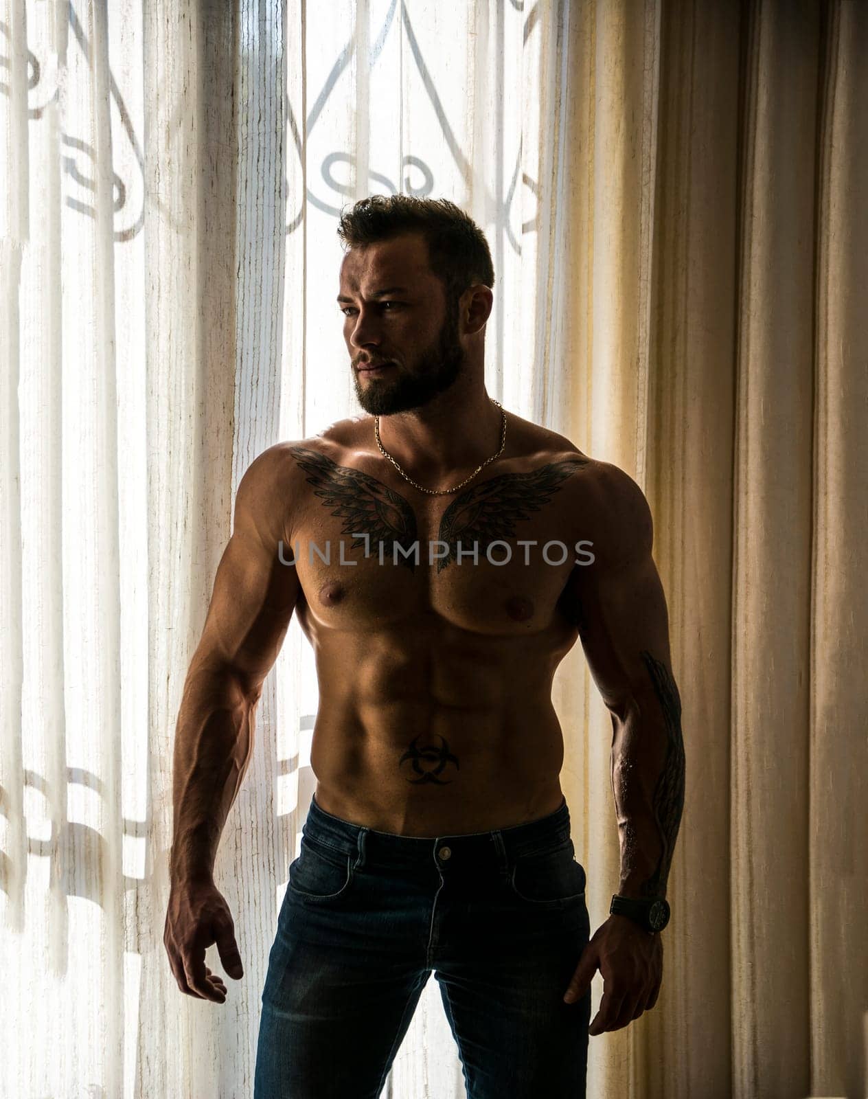 Portrait of sexy shirtless muscular man next to window curtains during the day, wearing only jeans