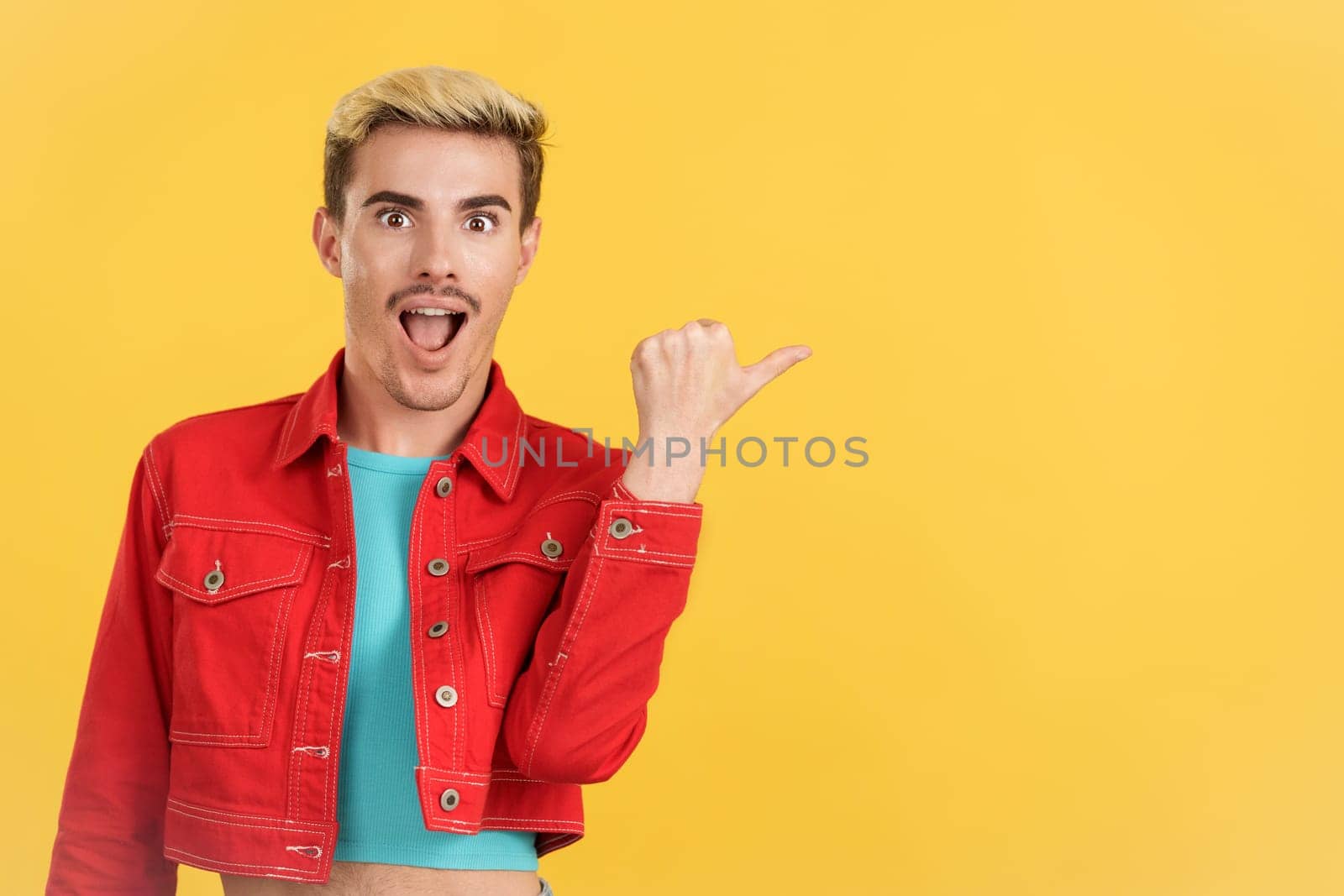 Surprised gay man pointing a blank space to the side in studio with yellow background