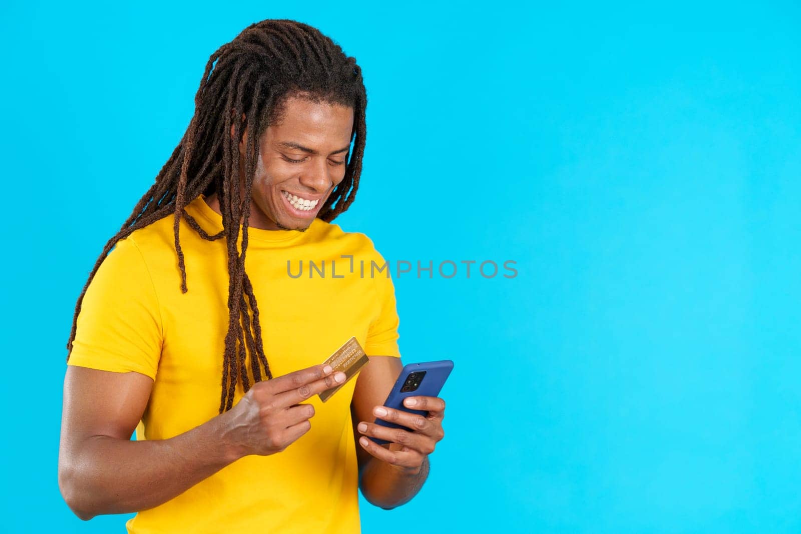 Happy latin man with dreadlocks using mobile and card to shopping online in studio with blue background