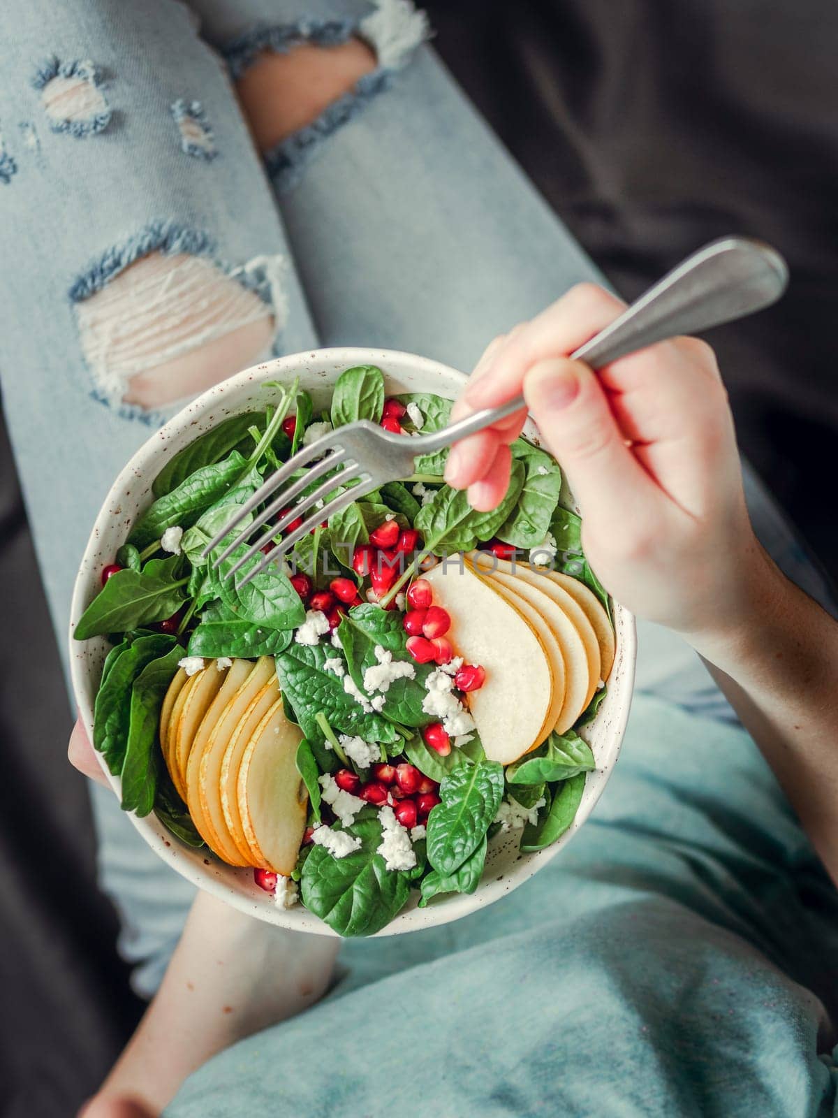Woman in jeans holding vegan salad bowl with spinach, pear, pomegranate, cheese. Vegan breakfast, vegetarian food, diet concept. Girl in jeans holding fork with knees and hands visible