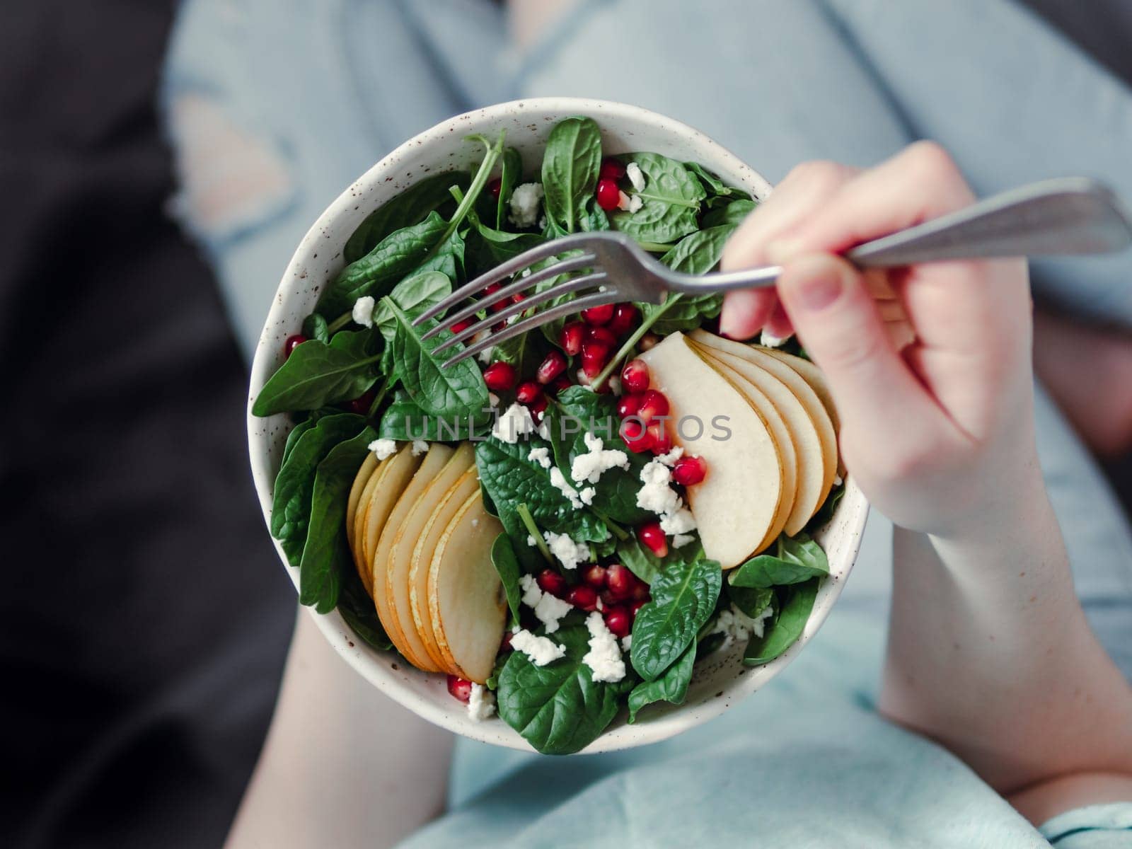 Woman in jeans holding vegan salad bowl with spinach, pear, pomegranate, cheese. Vegan breakfast, vegetarian food, diet concept. Girl in jeans holding fork with knees and hands visible
