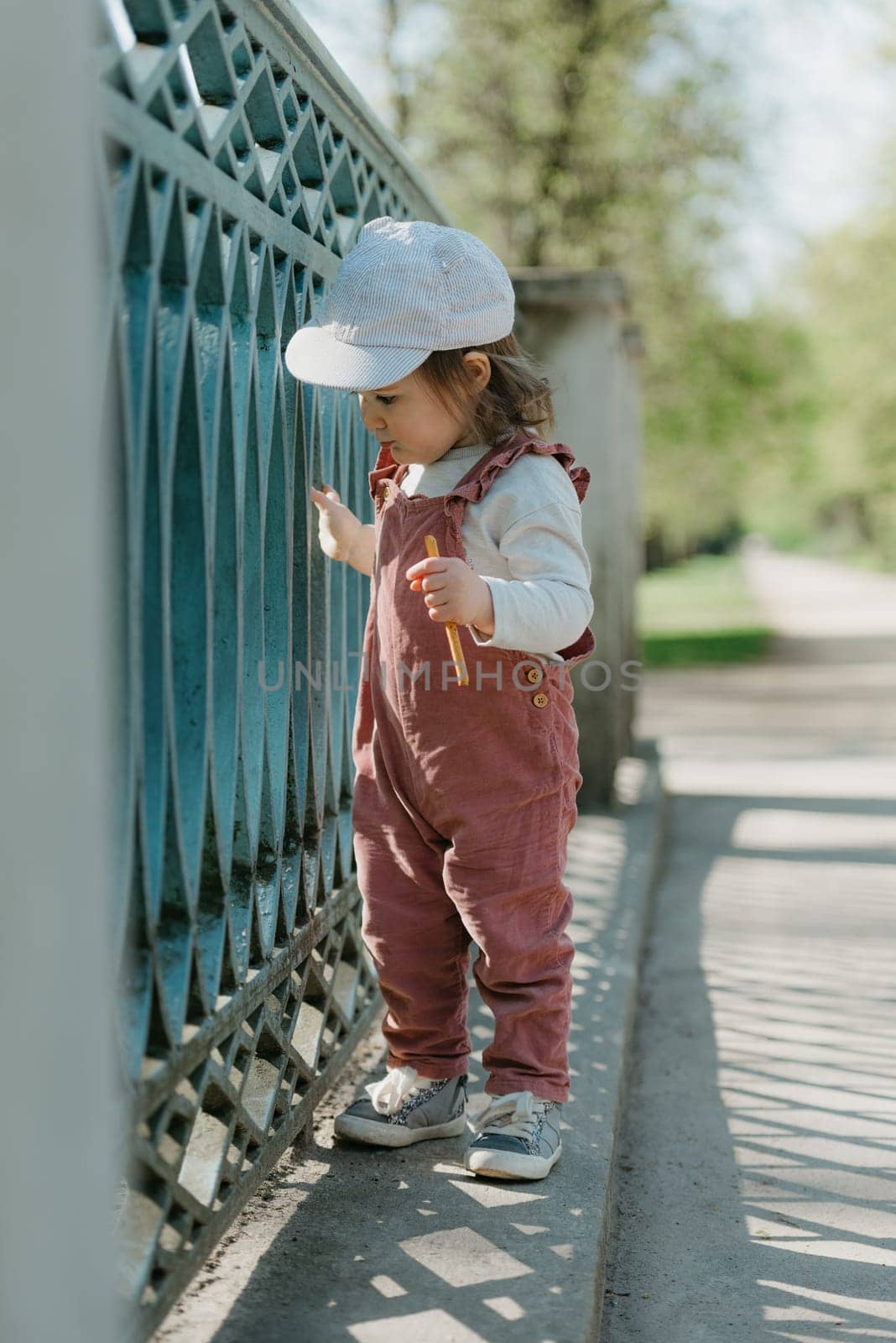 A female toddler eats a sausage close to the garden bridge fence. A baby girl in a cap and velvet overall staring to the side in the park.
