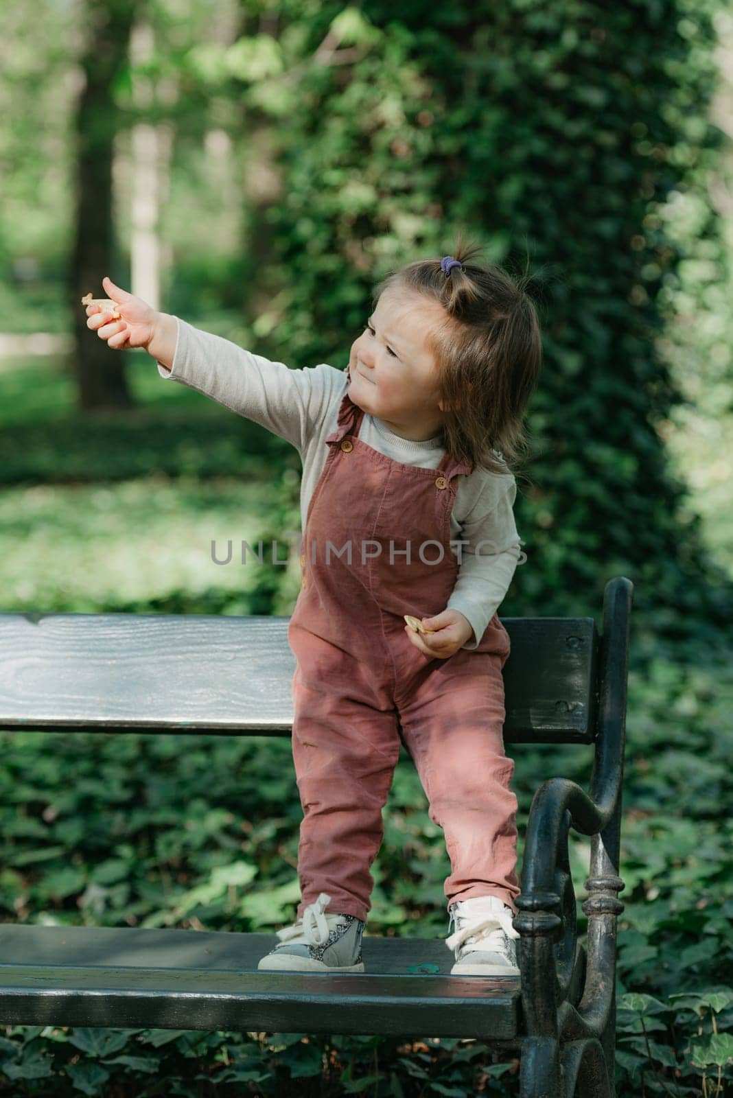 A female toddler in a velvet overall holds out a cookie on the garden bench. A baby girl with a small ponytail is having fun in the park.
