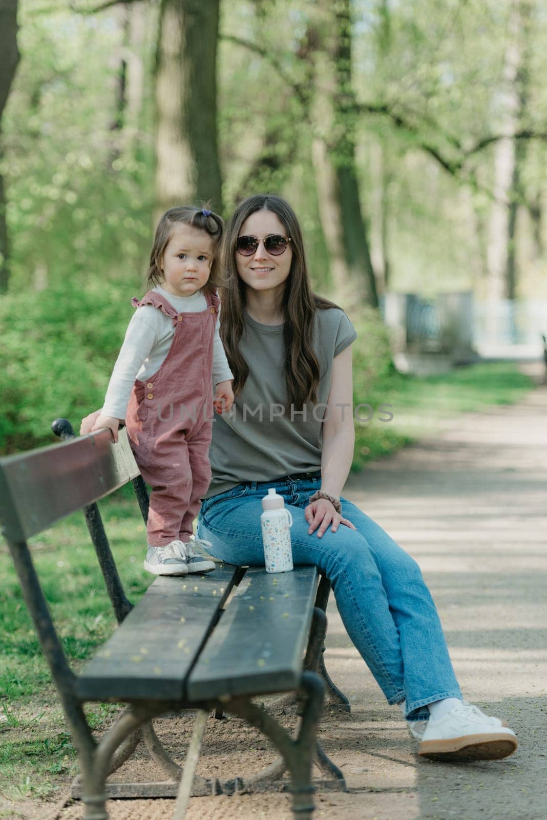 A mother sits close to her little daughter on the bench in the park. A female toddler in velvet overall is having fun with her mom in the garden.