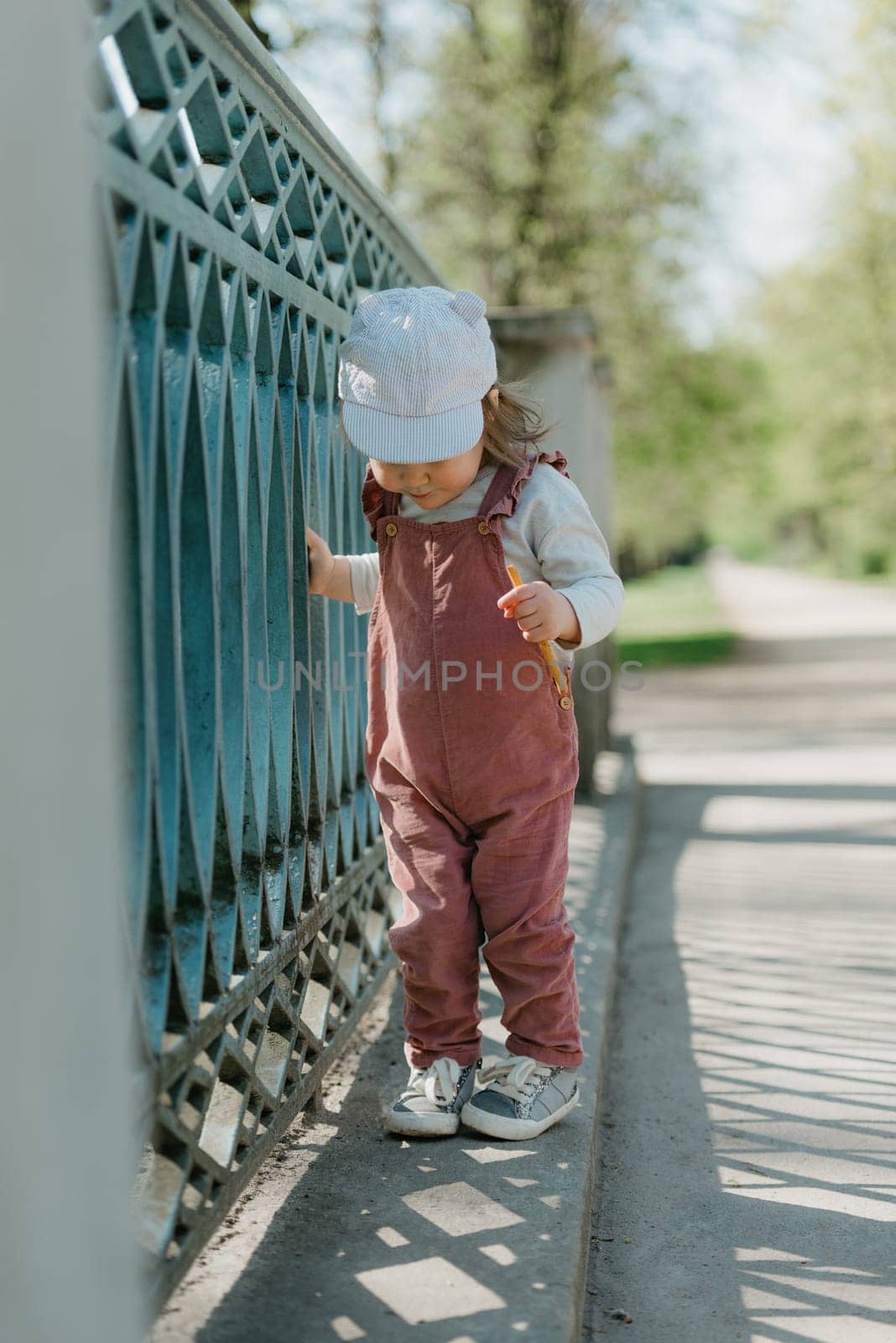 A female toddler eats a sausage close to the garden bridge fence. A baby girl in a cap and velvet overall walks in the park.