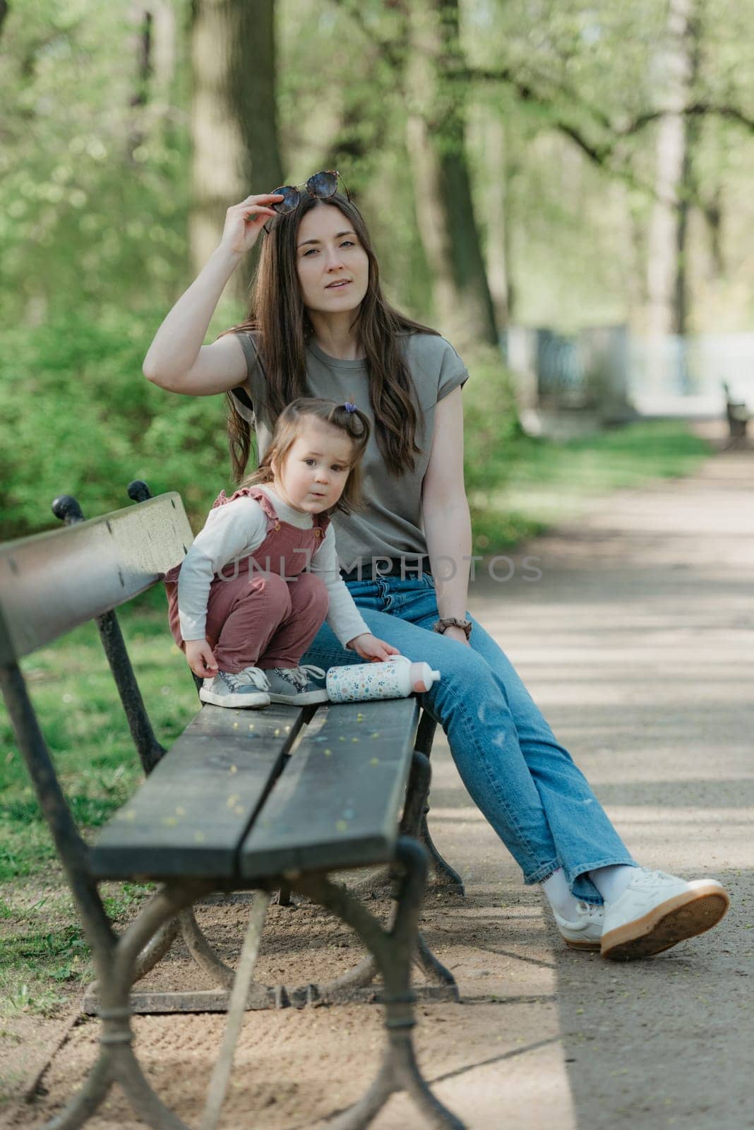 A mother rests close to her little daughter on the bench in the green park. A female toddler in velvet overall is playing with her mom in the garden.