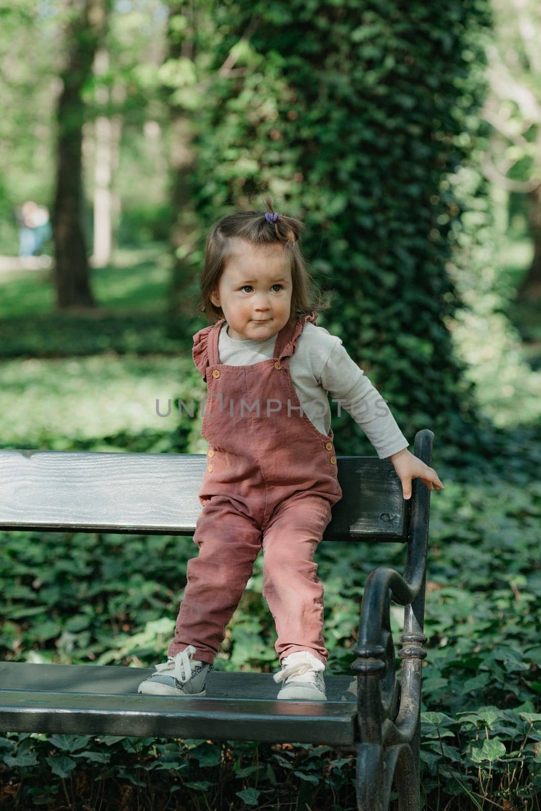 A female toddler in a velvet overall holds is on the garden bench. A baby girl with a small ponytail is having fun in the park.