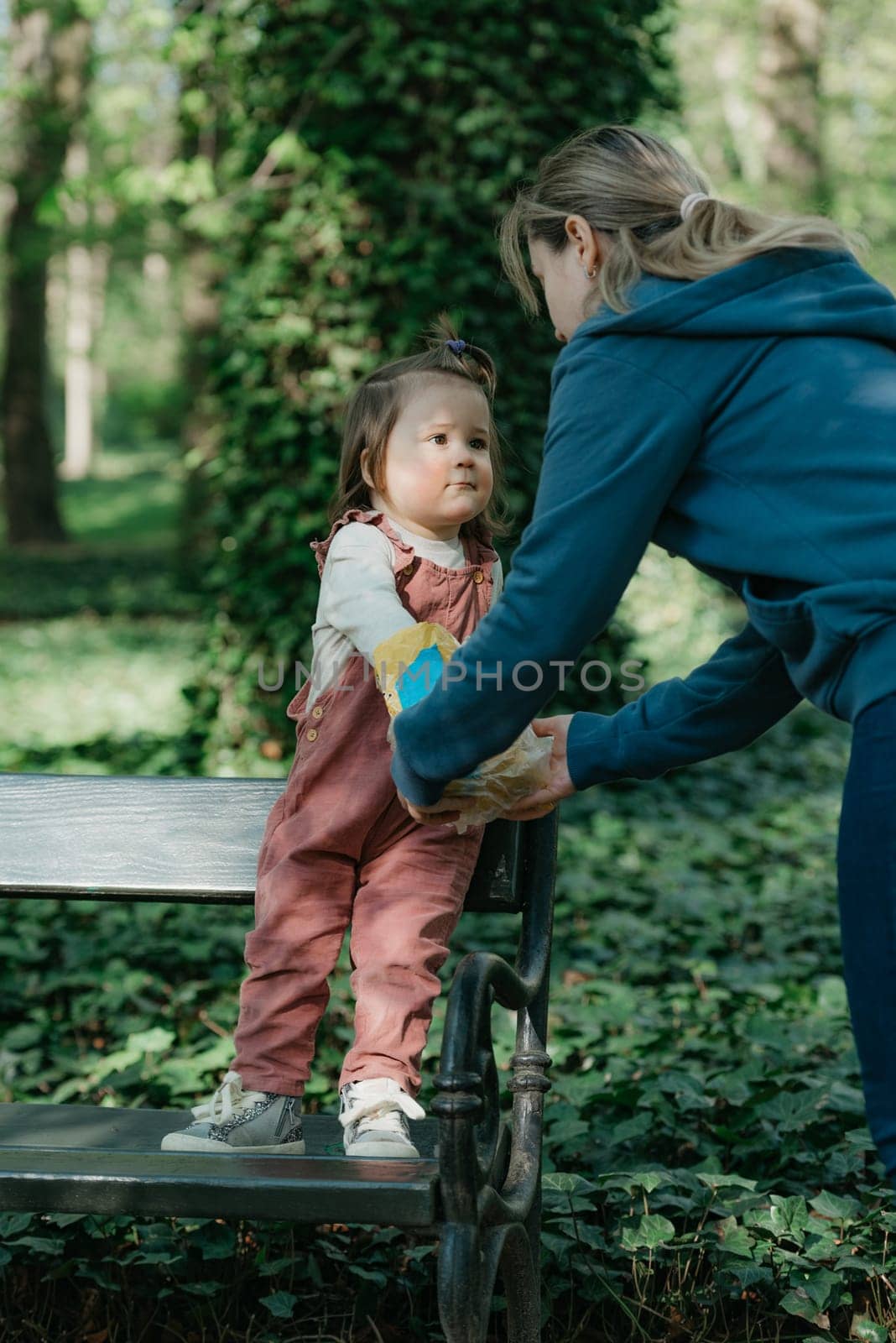 A mother with a ponytail feeds cookies to her little daughter in the park. A female toddler in velvet overall is having fun on the bench in the garden.