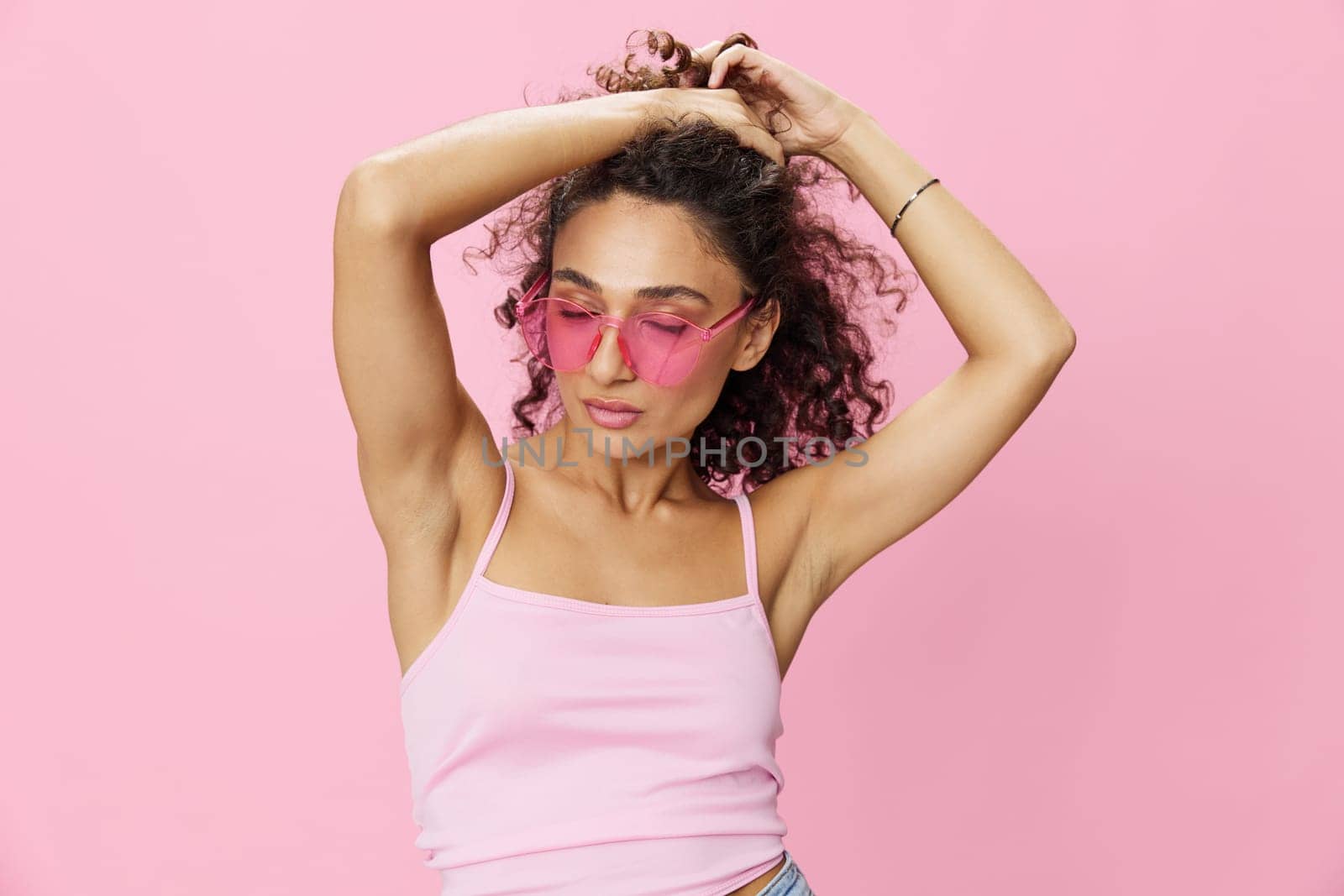 Happy woman hands up waxing her armpits, shaving, with curly hair in a pink tank top and jeans on a pink background wearing sunglasses with a nice tan, copy space. High quality photo