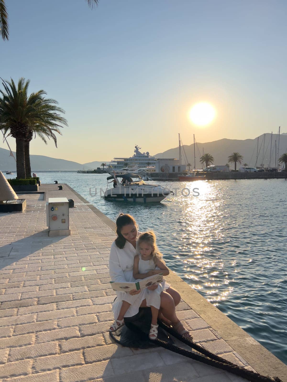 Mom reads a book to a little girl sitting on a bollard on a pier by the sea. High quality photo