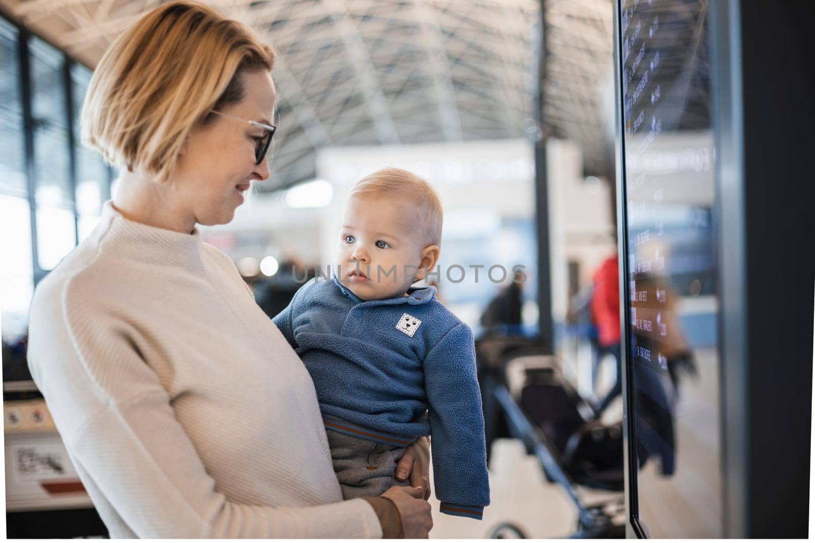 Mother traveling with child, holding his infant baby boy at airport terminal, checking flight schedule, waiting to board a plane. Travel with kids concept