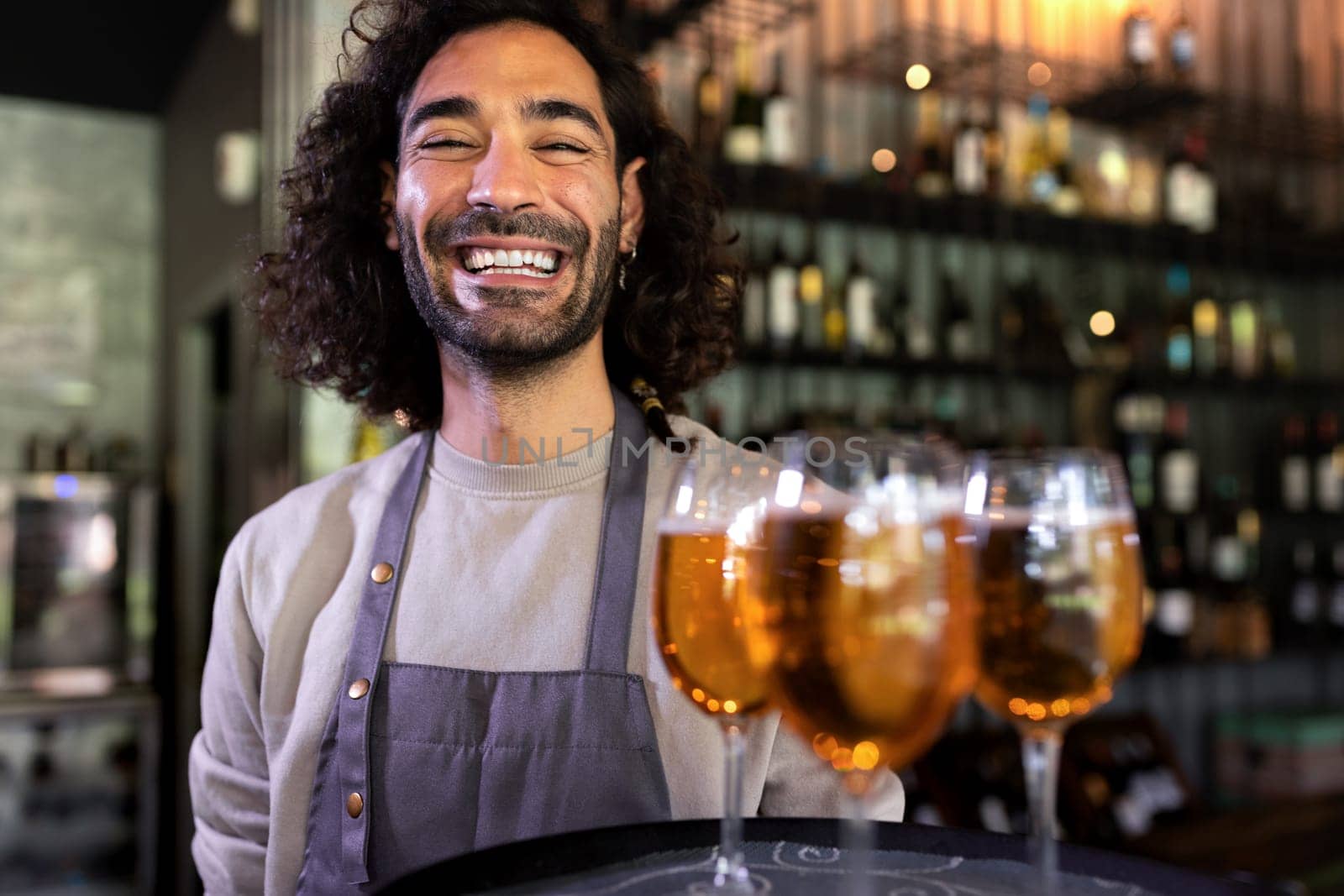Happy smiling young waiter wearing apron standing in bar holding tray of beers looking at camera. Restaurant concept.