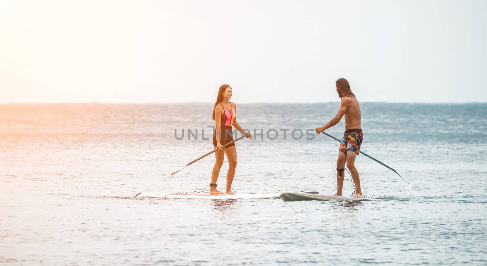 Sea woman and man on sup. Silhouette of happy young woman and man, surfing on SUP board, confident paddling through water surface. Idyllic sunset. Active lifestyle at sea or river